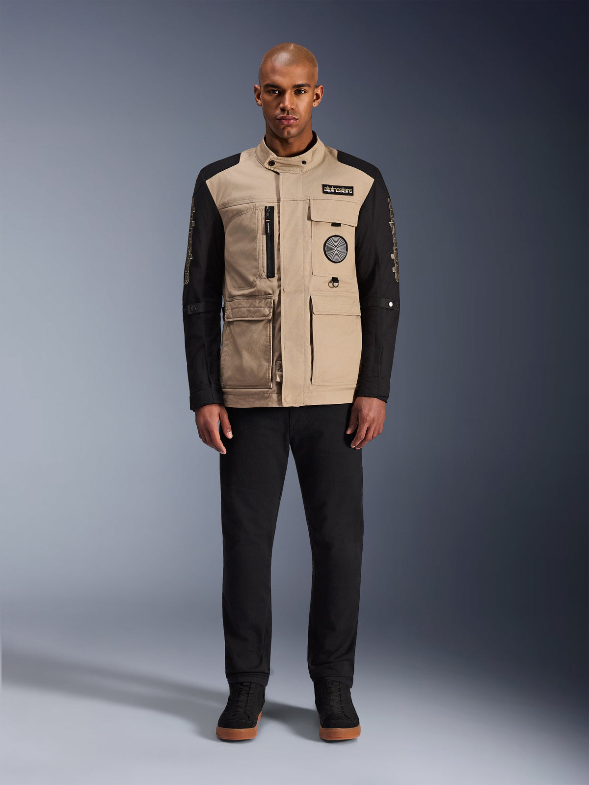 Alpinestars Dice-jakke, urban motorcykeljakke, aluminiums-tan krop med sorte ærmer, arbejdstøjsinspireret silhuet med fire funktionelle forlommer, bæres med sorte bukser og sorte high-top sneakers, robust og gadeklart køreudstyr