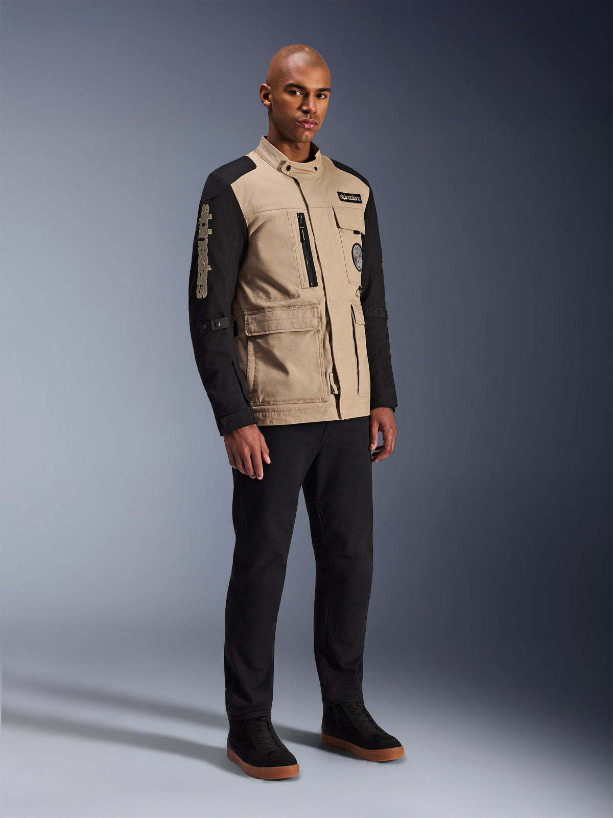 Alpinestars Dice-jakke, urban motorcykeljakke, Aluminum Black og tan, tofarvet design med sorte ærmer og khaki torso, båret af en model med sorte bukser og sorte sneakers, arbejdstøjsinspireret æstetik med funktionelle lommer og broderet branding på ærmet.
