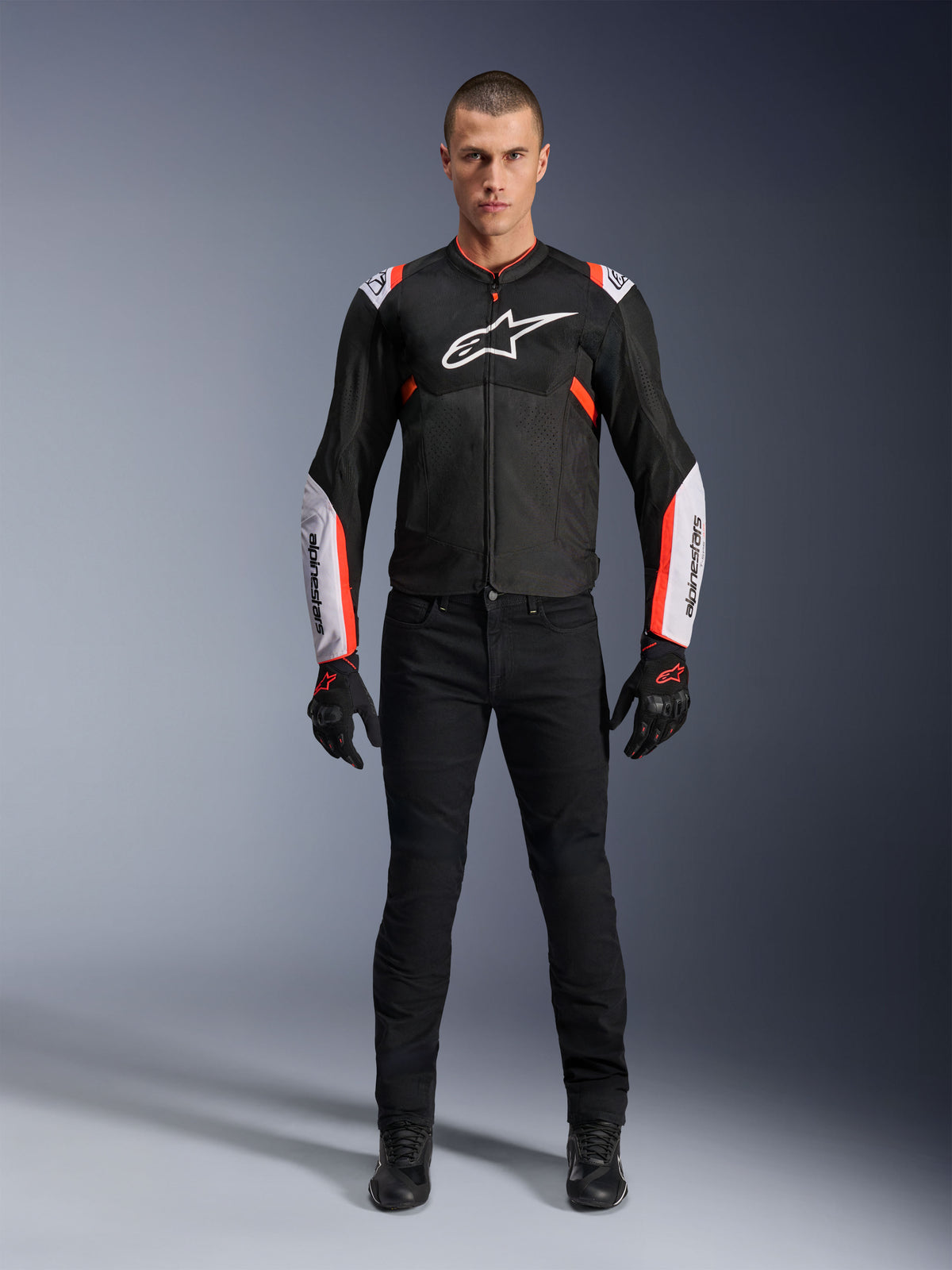 Male model wearing Alpinestars T-Sps Air V2 Jacket in Black White Red Fluo, featuring large tech mesh panels and bold white branding on the chest and sleeves, paired with black technical riding pants and gloves, urban sport motorcycle gear