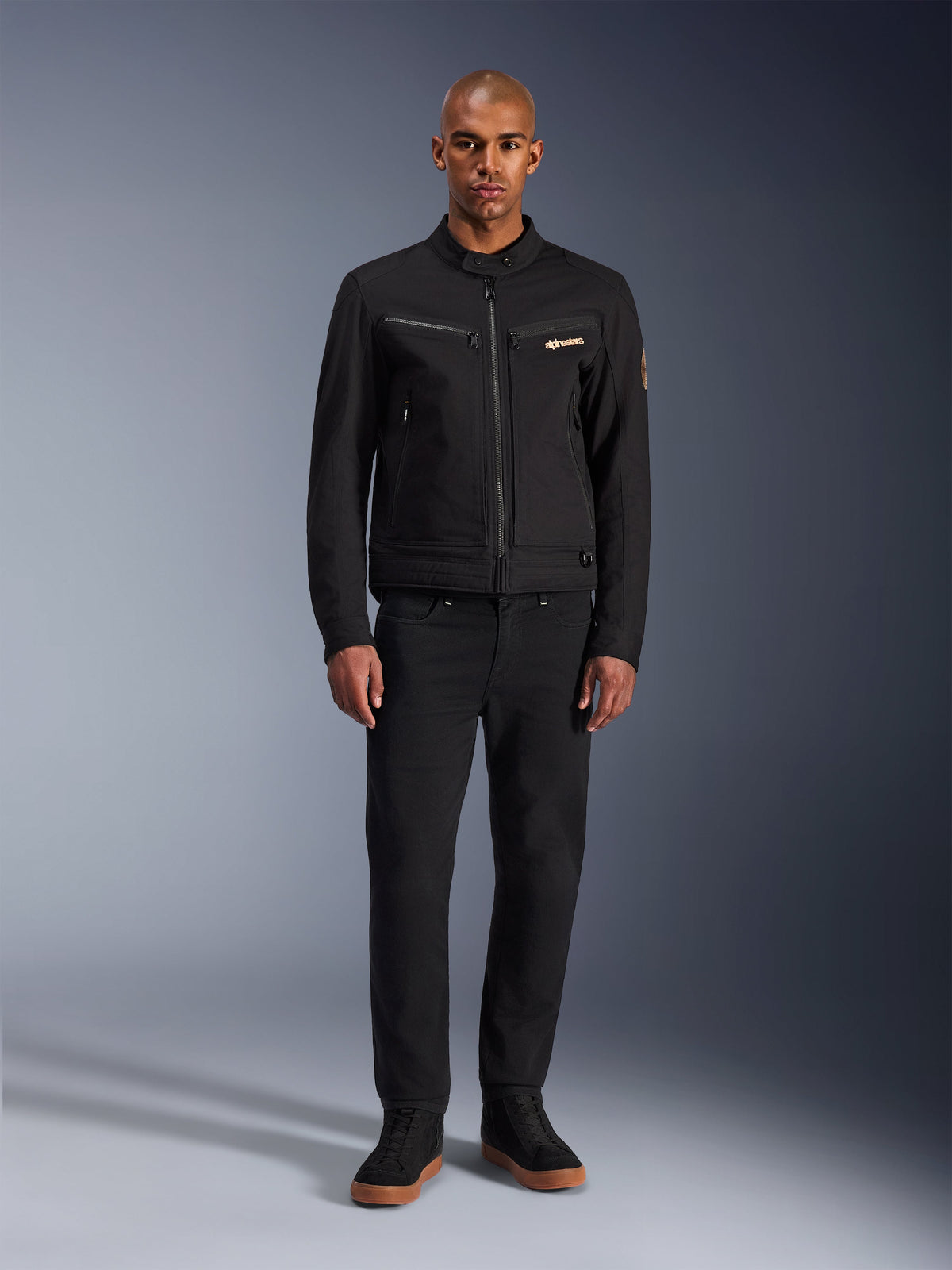 Alpinestars Bruiser jakke, sort urban motorcykeljakke, båret af model med sorte bukser og high-top sneakers, set forfra med brystlommer med lynlås og broderet logo, klassisk arbejdstøjsinspireret design til daglig pendling