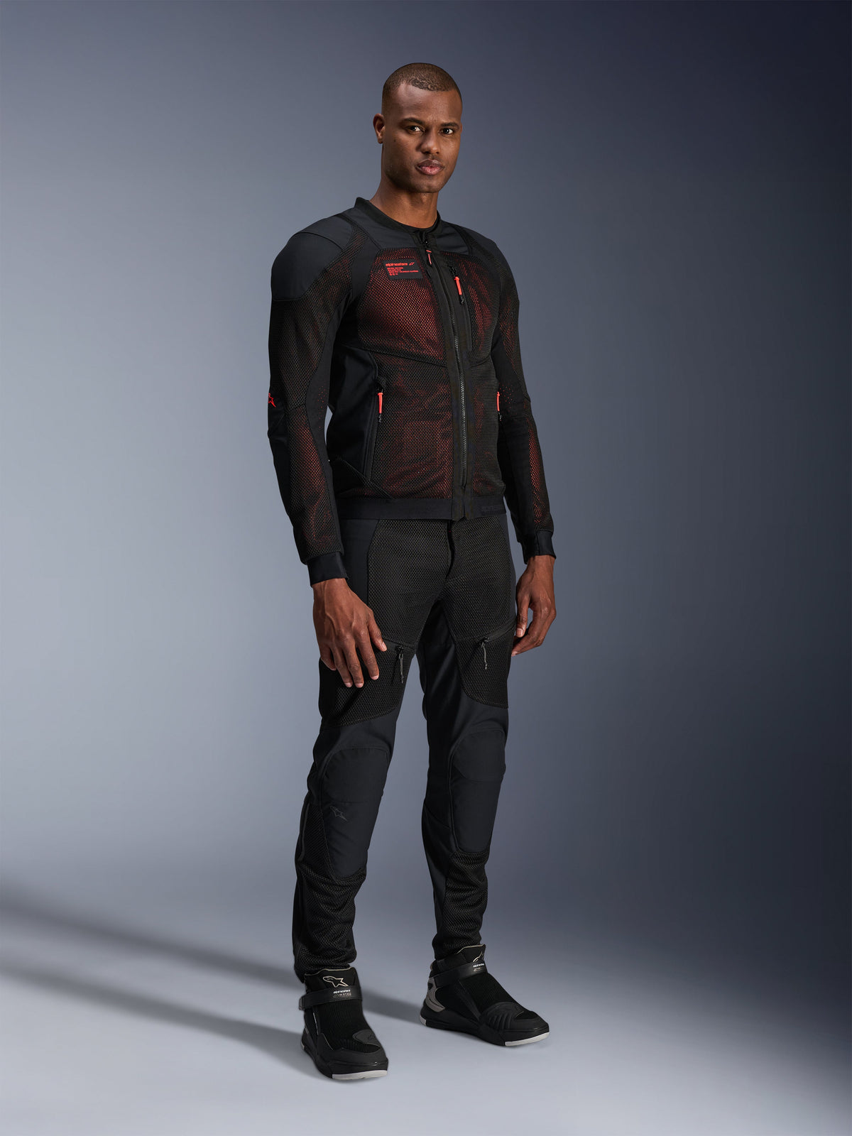 Alpinestars Troop-Air Pro Jakke i Black Red Fluo, teknisk motorcykeljakke i pansret mesh, båret af en mandlig model med matchende sorte pansrede bukser og high-top støvler, studieportræt i fuld længde.