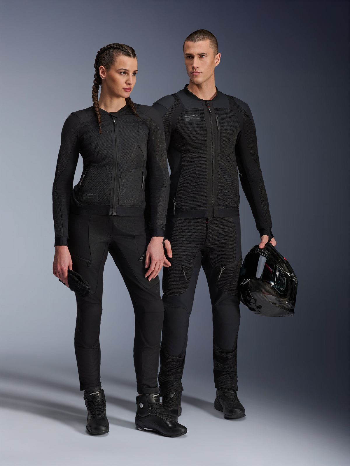 Alpinestars Stella Troop-Air jakke, mand og kvinde iført matchende sort teknisk motorcykeludstyr med mesh-konstruktion for luftgennemstrømning, sorte bukser og støvler, mand der holder en sort hjelm, urbant køreudstyr