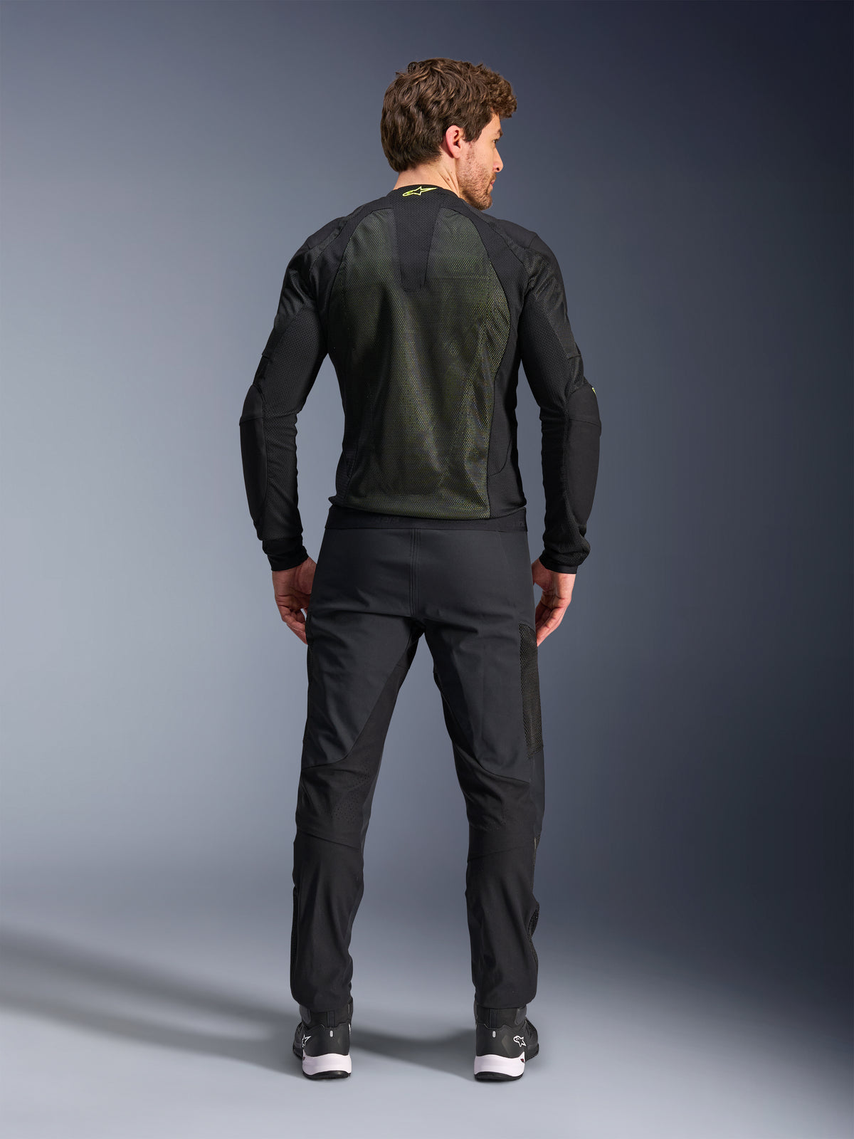 Alpinestars Troop-Air-jakke, sort fluorescerende gul, set bagfra på en model, med et stort 2D tech mesh-rygpanel til ventilation, teknisk motorcykeljakke båret med sorte kørebukser og støvler, urban motorcykelstil