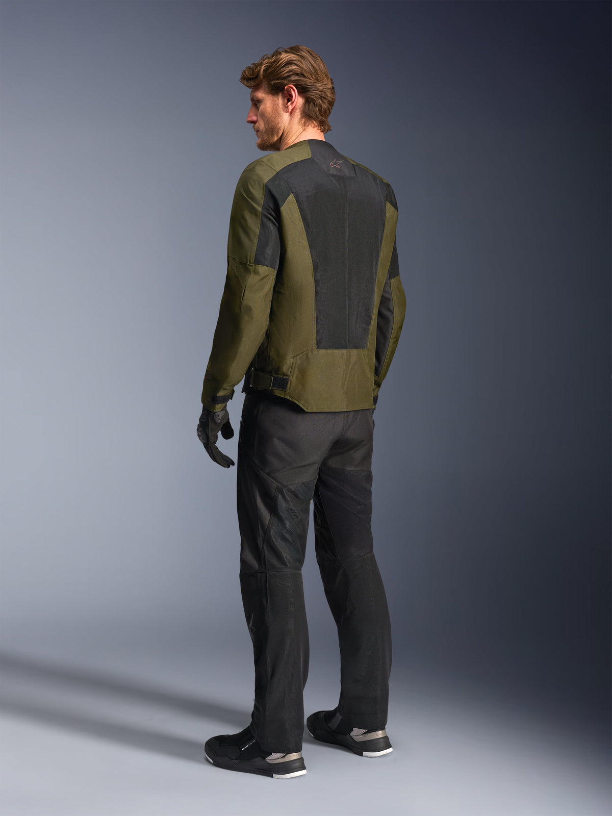 Mandlig model iført Alpinestars C-1 Air-jakke i Khaki, urban motorcykeljakke, set bagfra med fokus på store sorte mesh-paneler på ryggen, sorte tekniske kørebukser og handsker, moderne motorcykeludstyr til varmt vejr