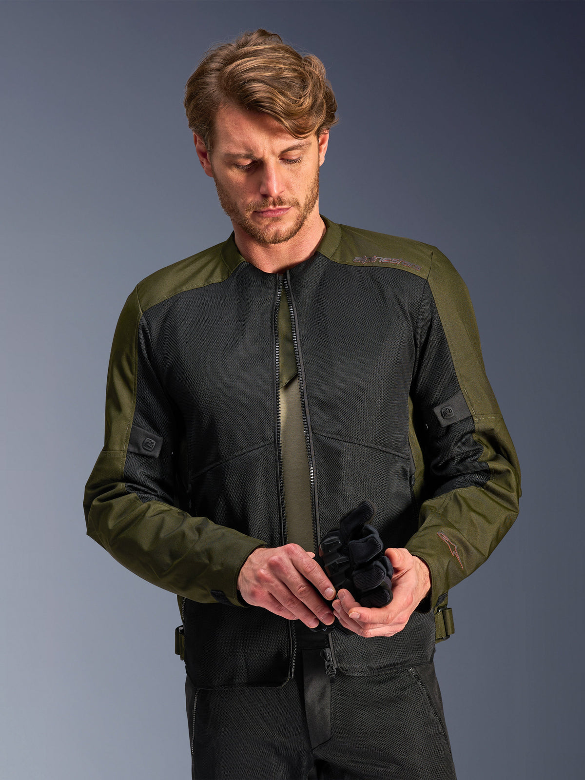 Mand iført Alpinestars C-1 Air-jakken i Khaki, urban motorcykeljakke med olivengrønt tekstil og sorte mesh-paneler, kigger ned mens han holder sorte motorcykelhandsker, rent og moderne design til landevejskørsel.