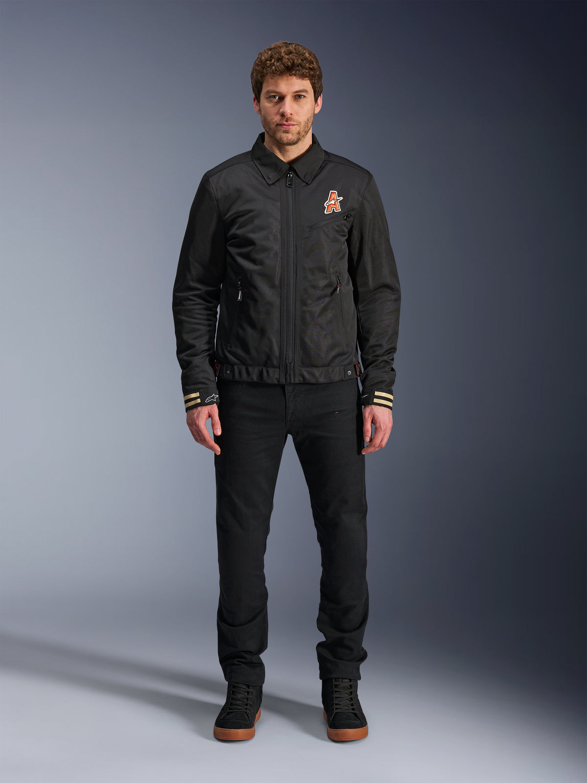 Male model wearing the Alpinestars Ignite Air Jacket in black, featuring a classic shirt collar, full front zipper, and an orange logo on the chest. Styled with black pants and black high-top sneakers for an urban contemporary motorcycle look.