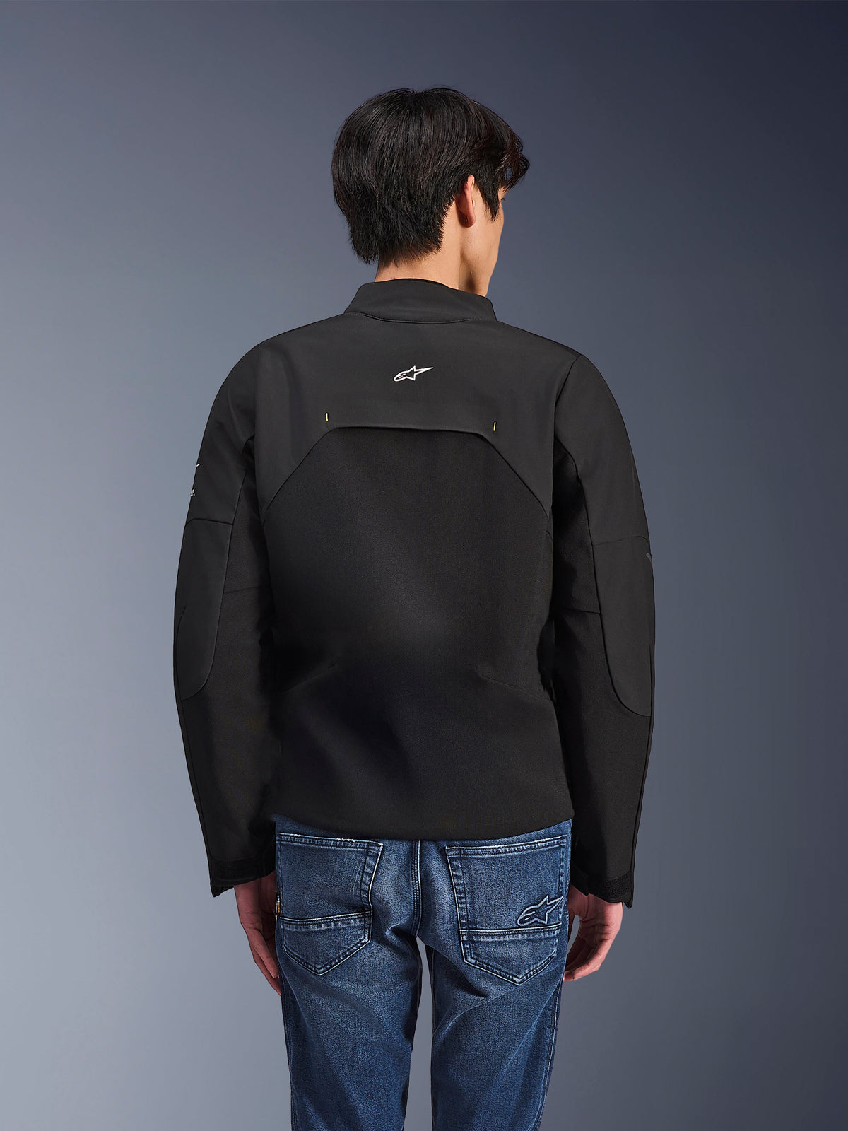 Alpinestars Provoke-jakke, urban motorcykeljakke, sort, set bagfra med hvidt Astars-logo og tekniske ripstop-paneler, båret med blå denimjeans, moderne design til pendling i byen