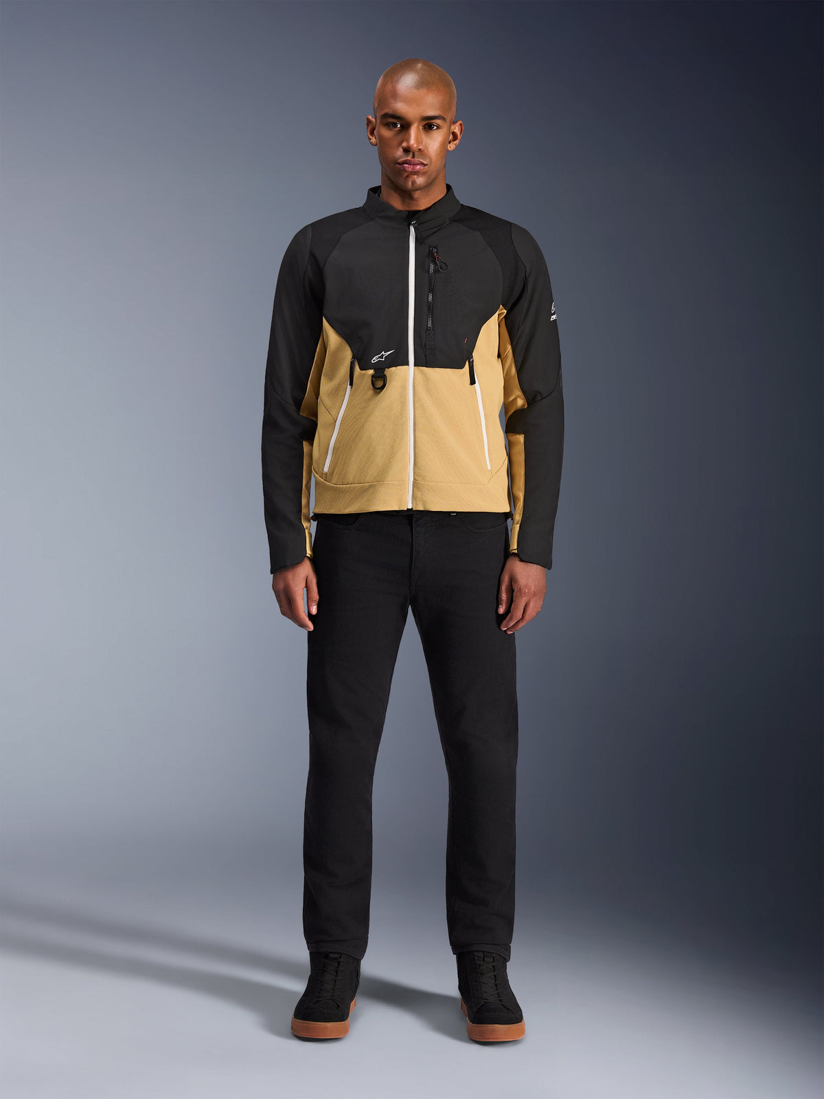 Alpinestars Provoke Jakke i Black Camel, urban motorcykeljakke med et tofarvet sort og camel design, båret af en model med sorte bukser og sorte sko, moderne silhuet med hvide lynlåse og teknisk softshell-konstruktion.