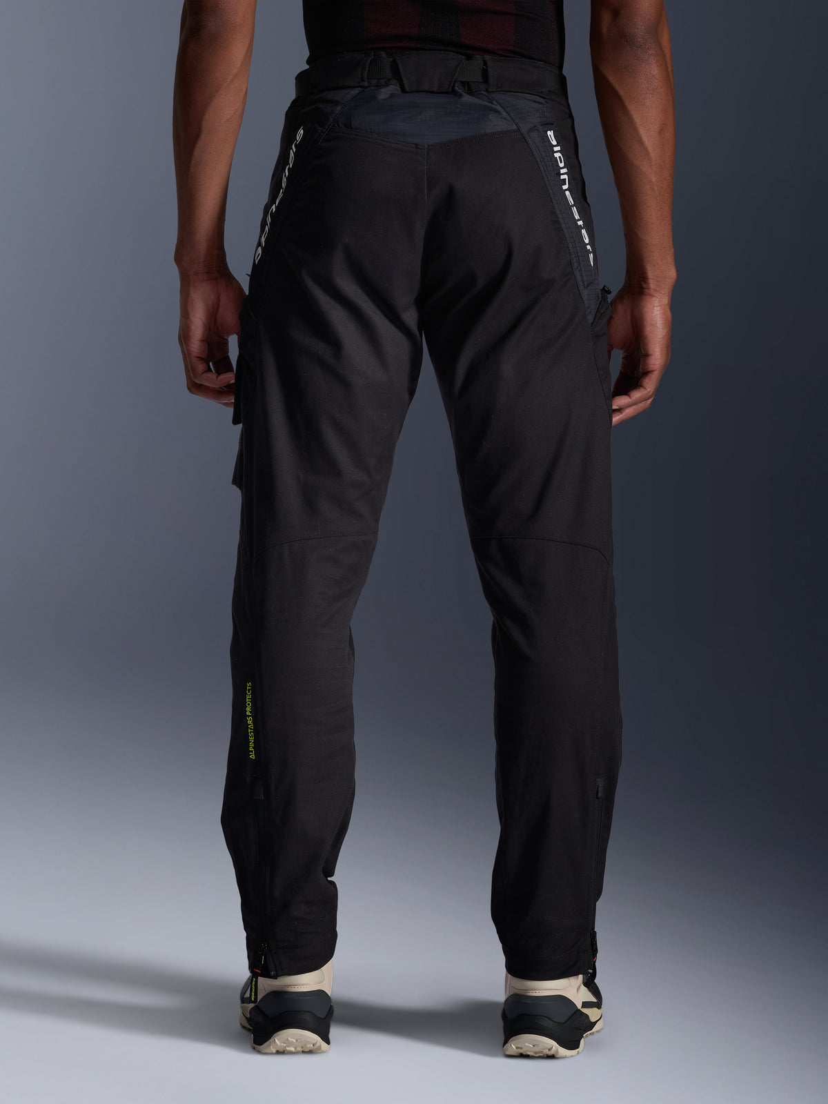 Alpinestars Acteon bukser, urban motorcykelbukser, sort, set bagfra med tilspidset pasform, hvid branding på de øvre ben og gult logo på den nederste del af læggen, justerbare benåbninger med lynlåse, bæres med cremefarvede sneakers