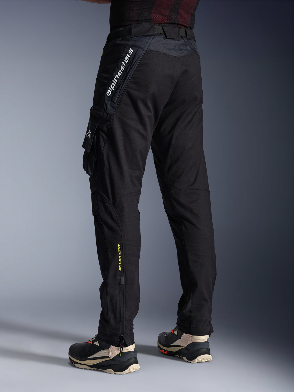 Alpinestars Acteon-bukser, sorte motorcykelbukser, set bagfra og fra siden med moderne tilspidset pasform, sidelomme med logo, lodret hvid Alpinestars-branding på låret, gul 'Alpinestars Protects'-tekst på underbenet, moderne urbant køreudstyr