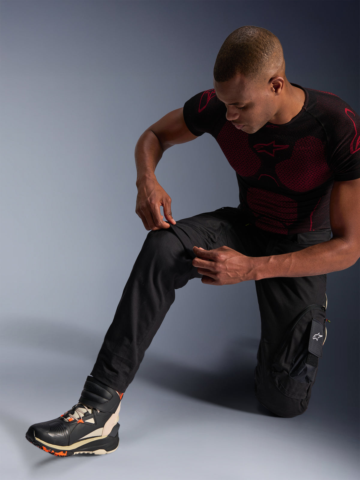 Alpinestars Acteon bukser, sorte urbane motorcykelbukser, båret af en knælende mand, der justerer knæområdet, med cargolommer og en tilspidset pasform, kombineret med et sort og rødt Alpinestars base layer og køresko