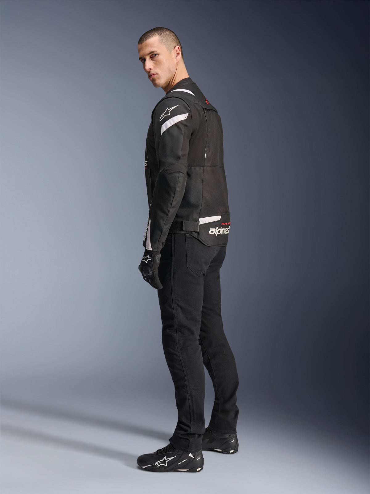 Alpinestars T-Stunt Air-jakke, sort og hvid, modulær motorcykeljakke med tech mesh-paneler, vist fra en profil fra siden og ryggen på en model, båret med sorte motorcykelbukser og sko, med hvide detaljer og branding på skuldre og ryg.