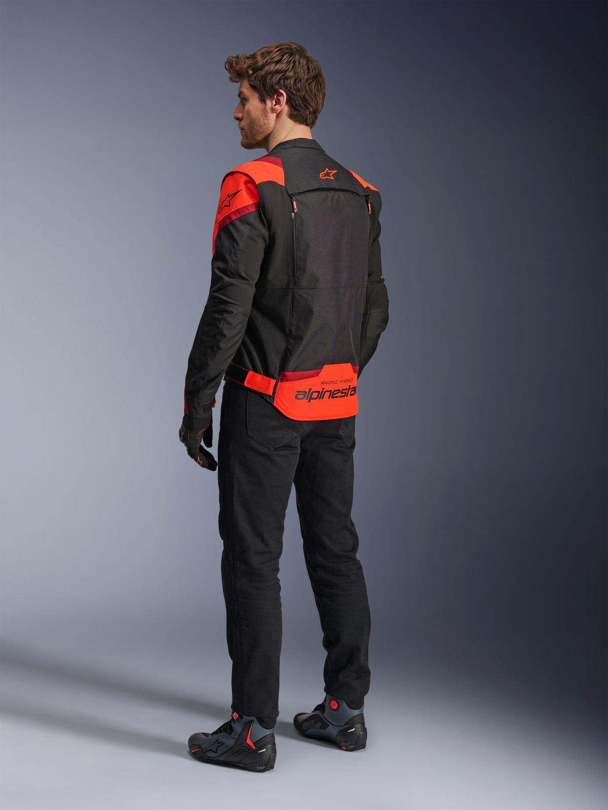 Man wearing Alpinestars T-Stunt Air Jacket from the back, Black Red Fluo and Dark Red, textile motorcycle jacket with mesh panels and sport-cut design, featuring bold Alpinestars branding on the lower back, styled with black pants and riding boots