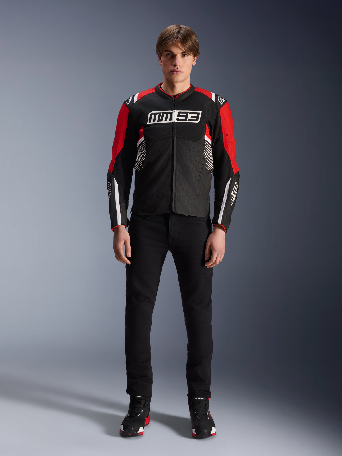 Mandlig model iført Alpinestars MM93 T-SPS Air V2-jakke i sort, klar rød og hvid, med Marc Márquez MM93-branding på brystet, mesh-paneler for øget luftgennemstrømning, kombineret med sorte bukser og sneakers i et studiemiljø.