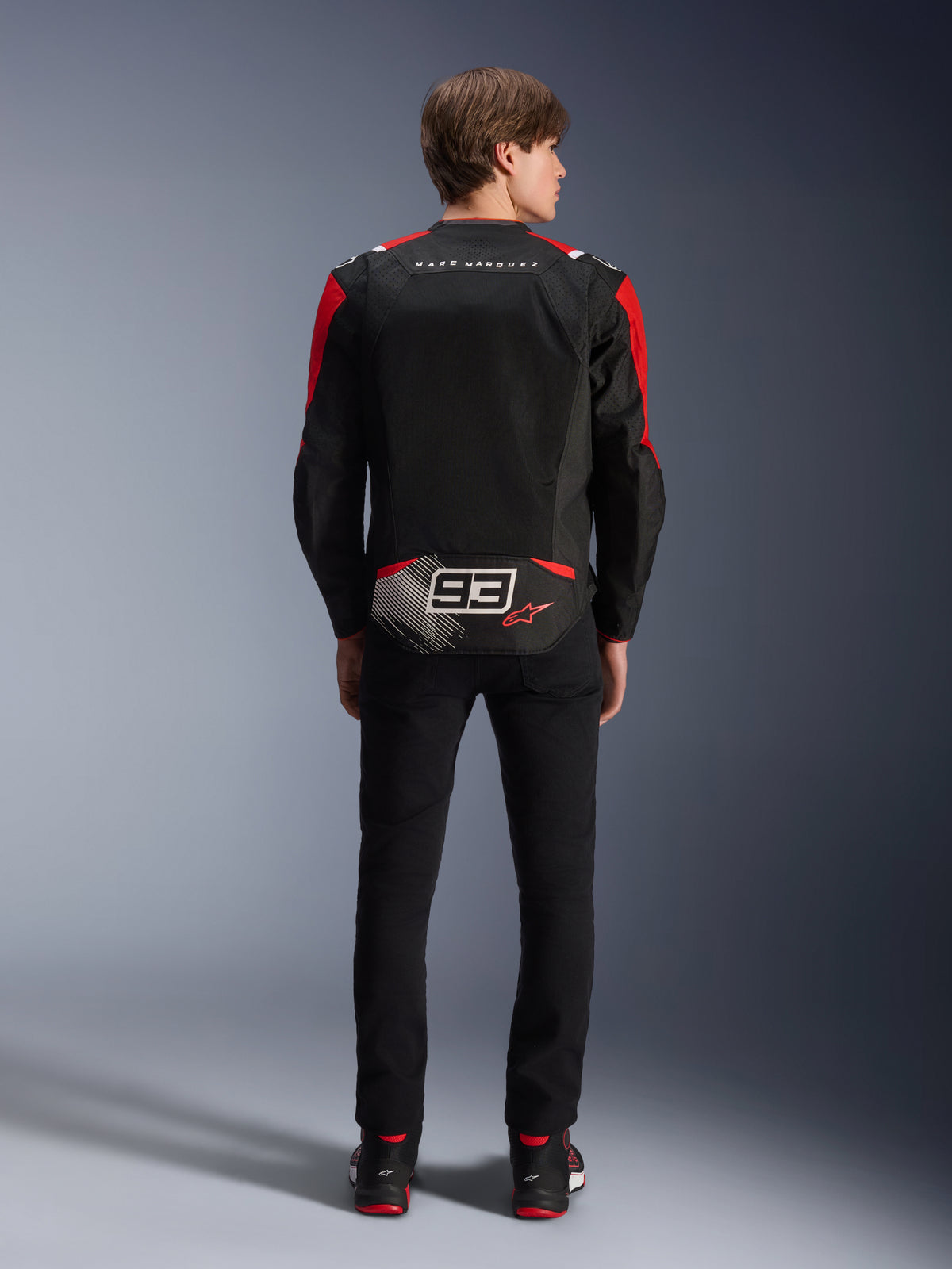 Alpinestars MM93 T-SPS Air V2-jakke, set bagfra på model, sort med klare røde og hvide detaljer, med Marc Marquez-signaturbranding og 93-logo på ryggen, performance-tekstilmotorcykeljakke til bykørsel.