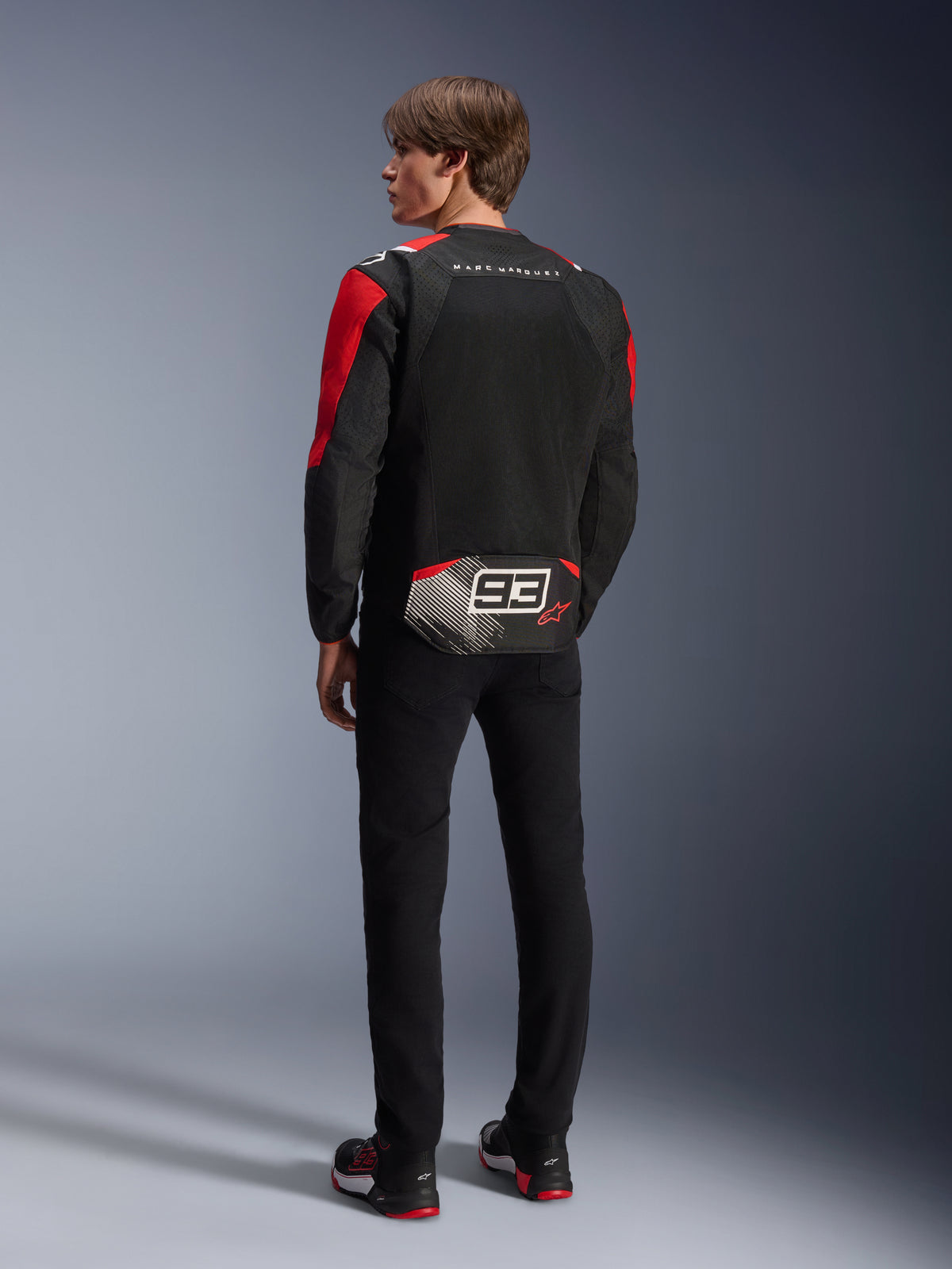 Model bagfra iført Alpinestars MM93 T-SPS Air V2-jakke, sort med klare røde og hvide detaljer, 'MARC MARQUEZ'-tekst og stort '93'-logo på lænden, mesh-tekstilkonstruktion til ventilation, kombineret med sorte bukser og matchende sort-røde kørersko