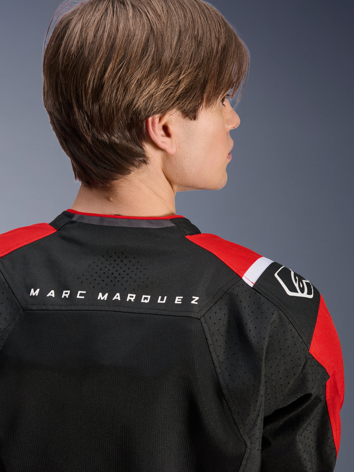 Alpinestars MM93 T-SPS Air V2 jakke, set bagfra af den øverste del af ryggen og skuldrene, sort med klare røde og hvide detaljer, 'MARC MARQUEZ' tekst over bærestykket, med tekniske mesh-paneler og skulderlogo, urban sport motorcykelbeklædning