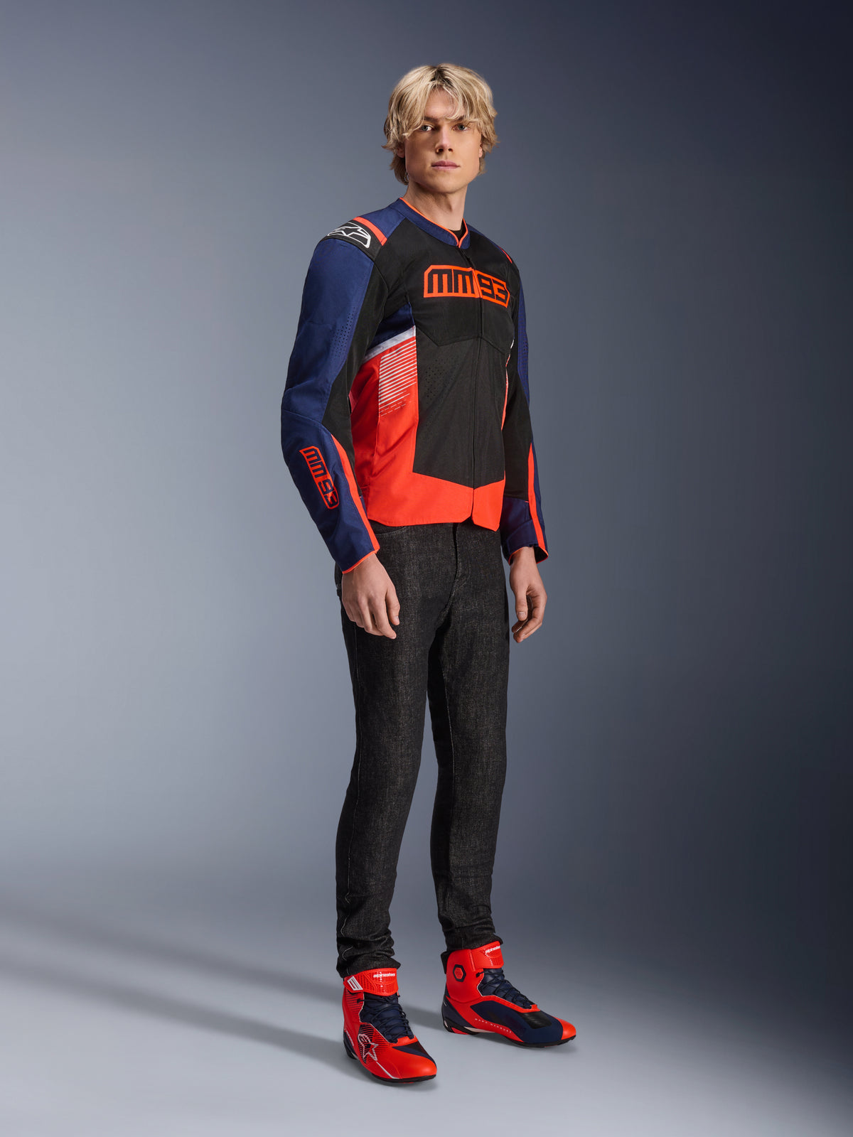 Mandlig model iført Alpinestars MM93 T-SPS Air V2-jakke i Blue Navy, sort og Red Fluo, med markant MM93-branding på brystet og ærmerne, kombineret med mørke jeans og matchende røde og blå motorcykelstøvler, sporty urbant køreudstyr