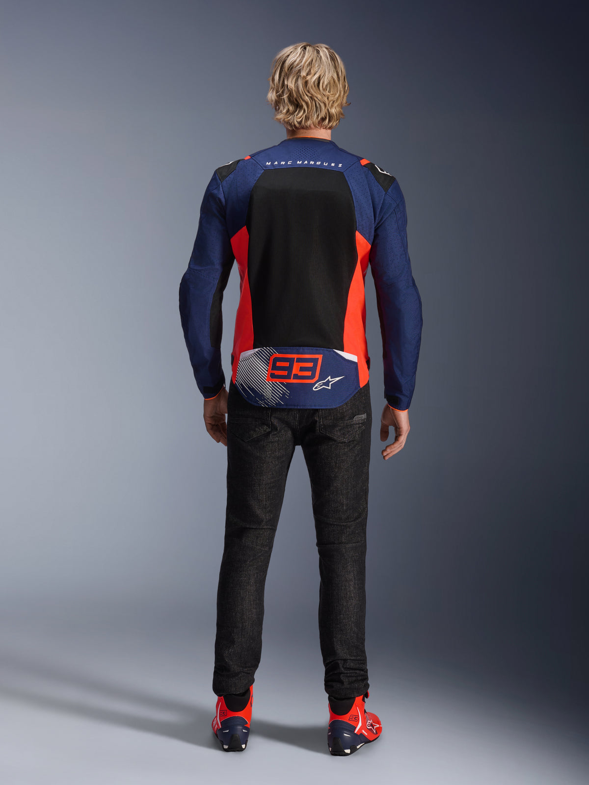 Bagfra af en model iført MM93 T-SPS Air V2-jakken i Blue Navy med Red Fluo og sorte detaljer, med et stort mesh-rygpanel og '93' Marc Marquez-branding, kombineret med mørke denimbukser og matchende kørestøvler.