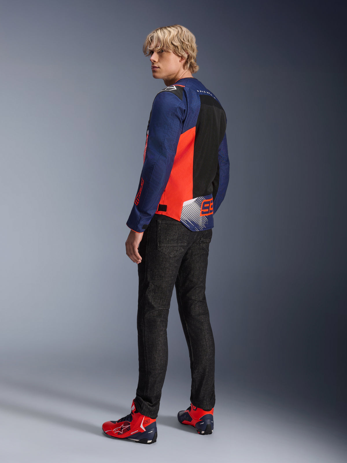 Alpinestars MM93 T-SPS Air V2 Jakke, mørkeblå og fluorescerende rød, vist bagfra på en model med sorte kørejeans og matchende røde og blå sko, med mesh-paneler og Marc Márquez 93-branding.