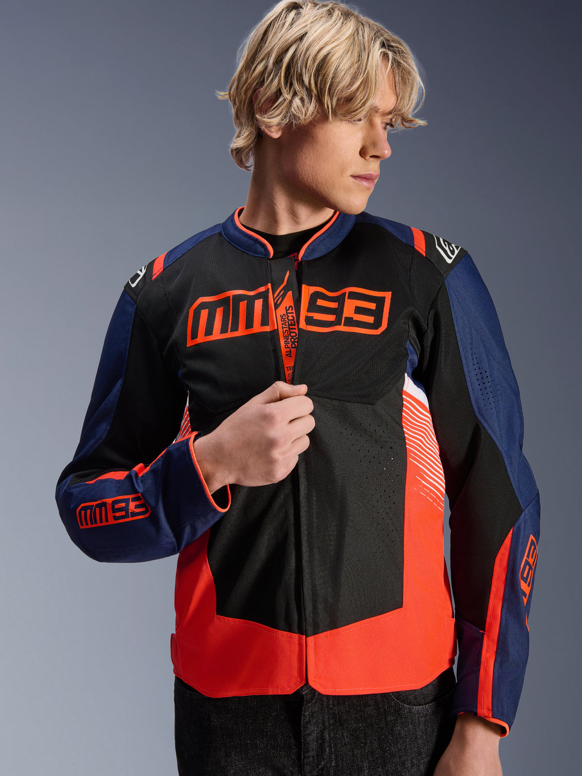 Alpinestars MM93 T-SPS Air V2 Jakke, mørkeblå, sort og fluorescerende rød tekstil motorcykeljakke, båret af en mandlig model, der justerer lynlåsen, med markant Marc Márquez MM93-branding på brystet og ærmerne, ventilerede mesh-paneler til sommerkørsel på landevej.