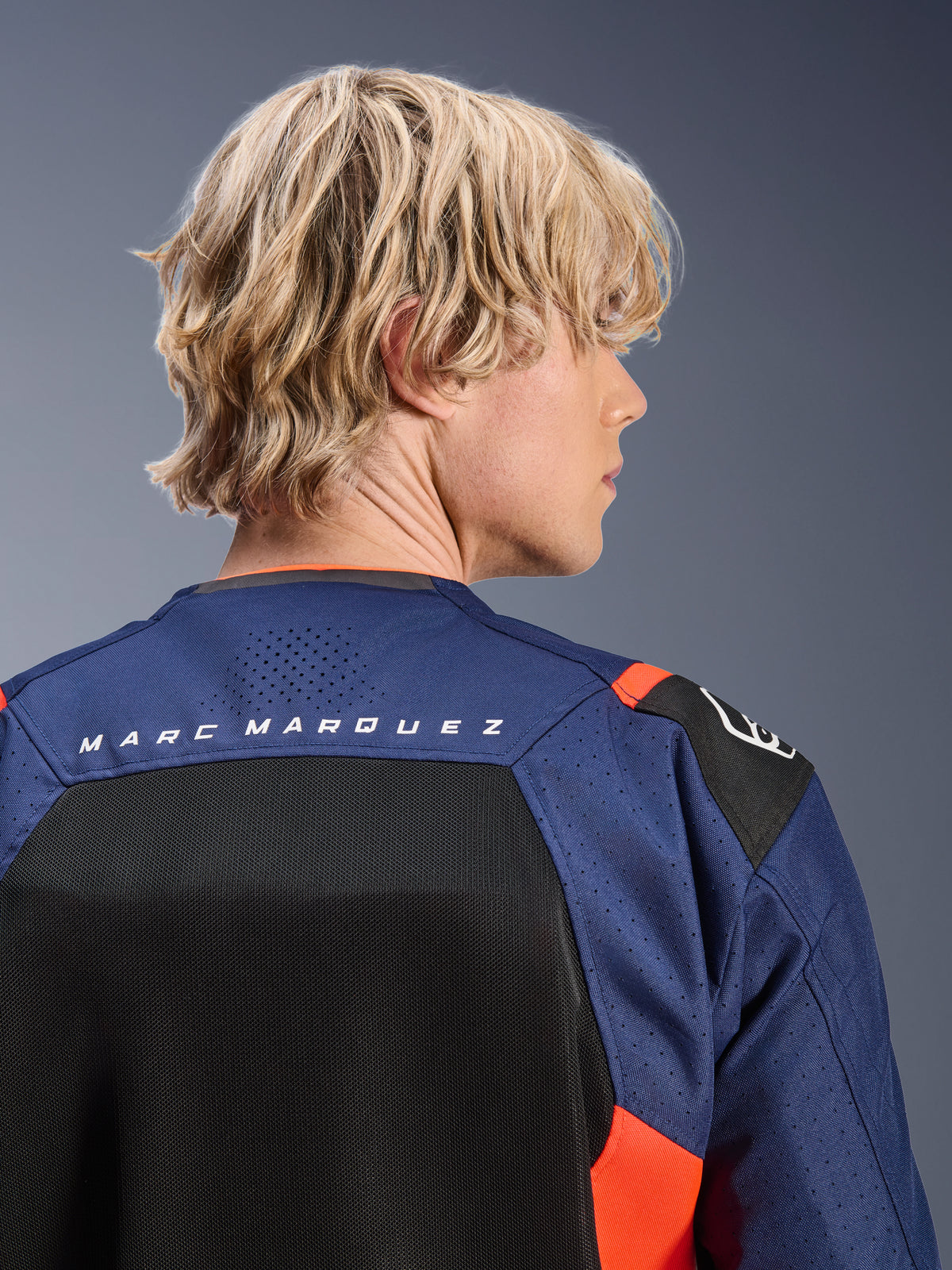 Alpinestars MM93 T-SPS Air V2-jakke, Blue Navy og sort med røde fluo-detaljer, nærbillede af den øverste del af ryggen med 'MARC MARQUEZ'-branding, åndbare mesh-paneler og perforeret tekstilkonstruktion, båret af en lyshåret model.