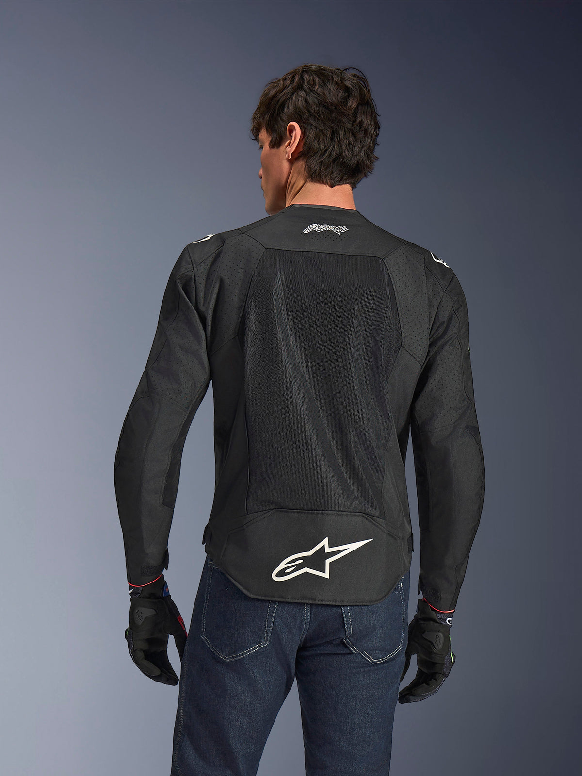 Alpinestars FQ20 T-SPS Air V2 Monster-jakke, sort, set bagfra med store ventilationspaneler i mesh og hvidt Alpinestars-logo nederst på ryggen, båret med blå jeans og sorte motorcykelhandsker, urban sport-kørestil.