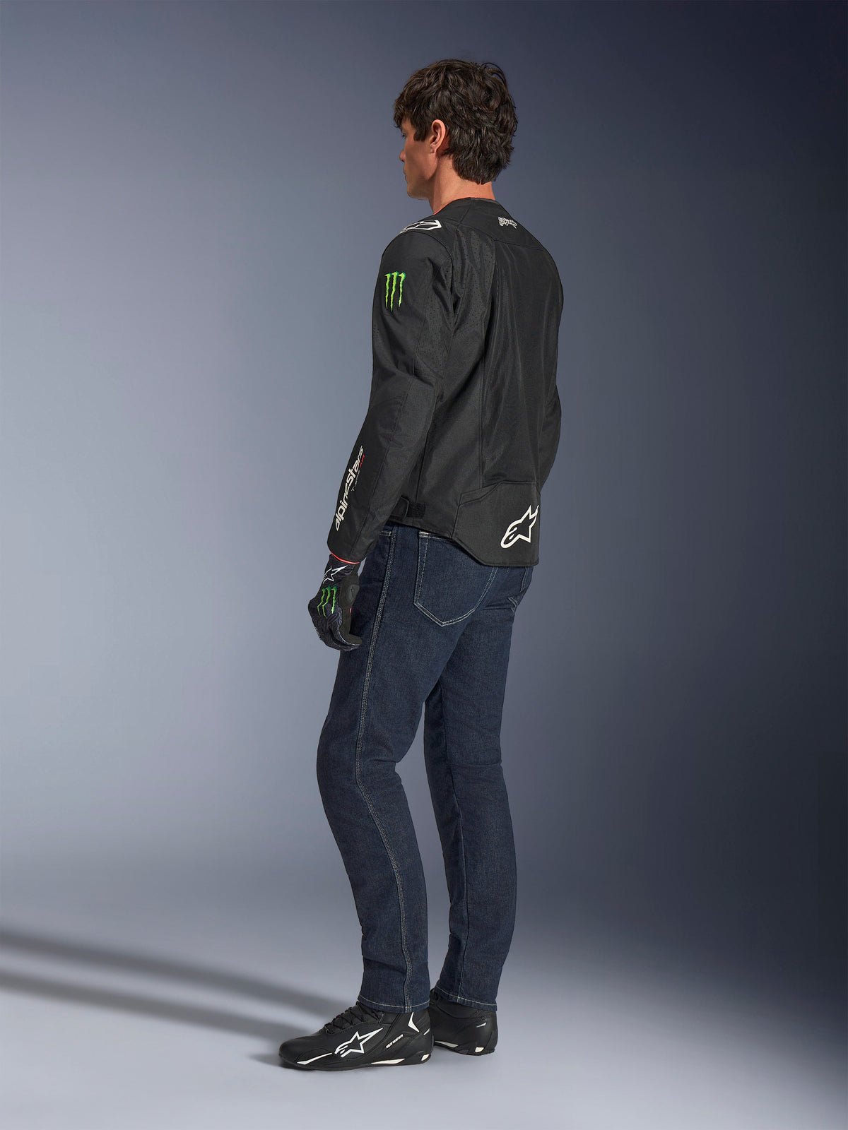 Alpinestars FQ20 T-SPS Air V2 Monster-jakke, sort tekstil-motorcykeljakke med grønne Monster Energy-logoer på ærmerne og hvidt Alpinestars-branding, vist fra en trekvart vinkel bagfra på en model med mørkeblå jeans og sorte køresko, urban sportsstil.