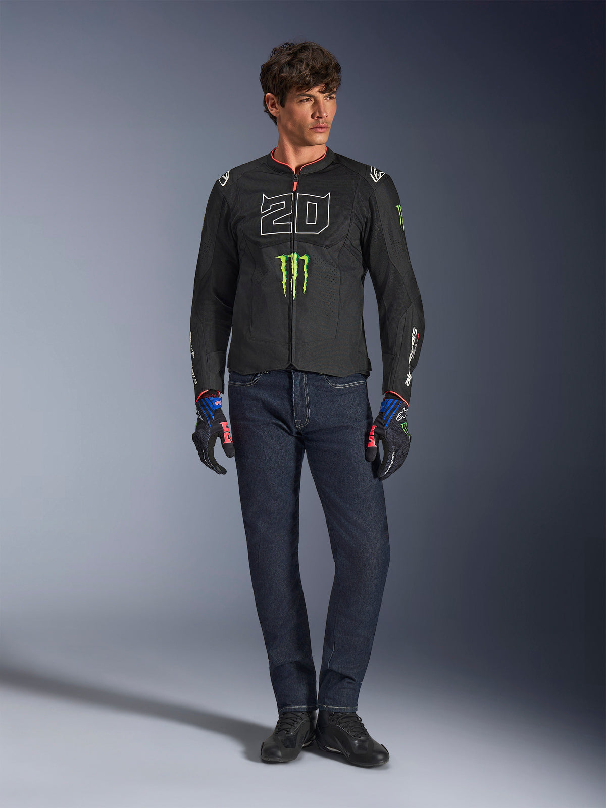 Mandlig motorcyklist iført Alpinestars FQ20 T-SPS Air V2 Monster-jakke, sort med grønt Monster Energy-logo og hvid '20'-grafik på brystet, kombineret med mørkeblå jeans og tekniske kørehandsker, urbant sport-motorcykeloutfit.
