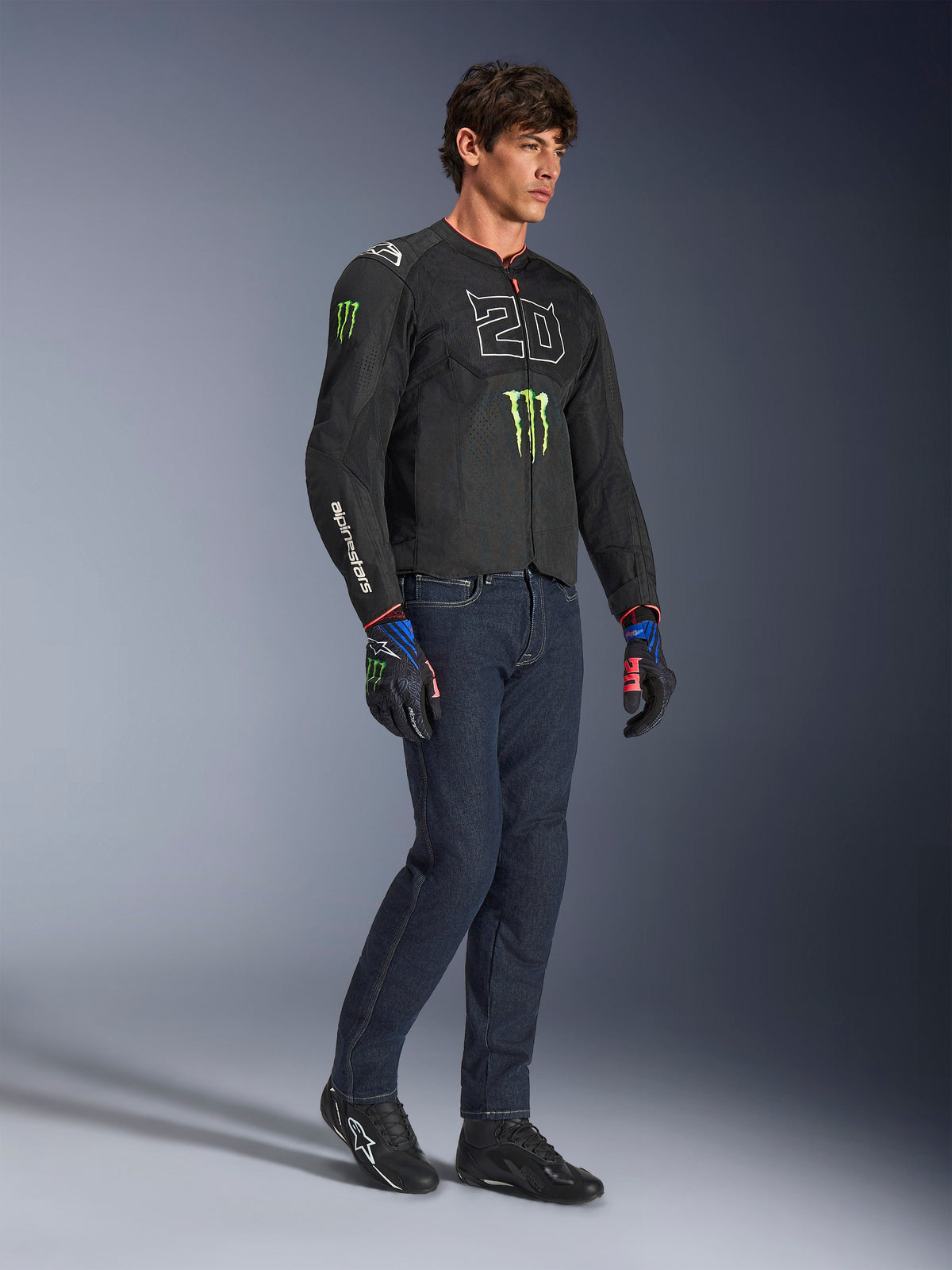 Mandlig motorcyklist iført Alpinestars FQ20 T-SPS Air V2 Monster-jakke i sort, med '20'-logo og Monster Energy-klo på brystet, kombineret med mørke motorcykeljeans, sorte motorcykelsko og matchende FQ20-handsker, studieoptagelse i fuld figur