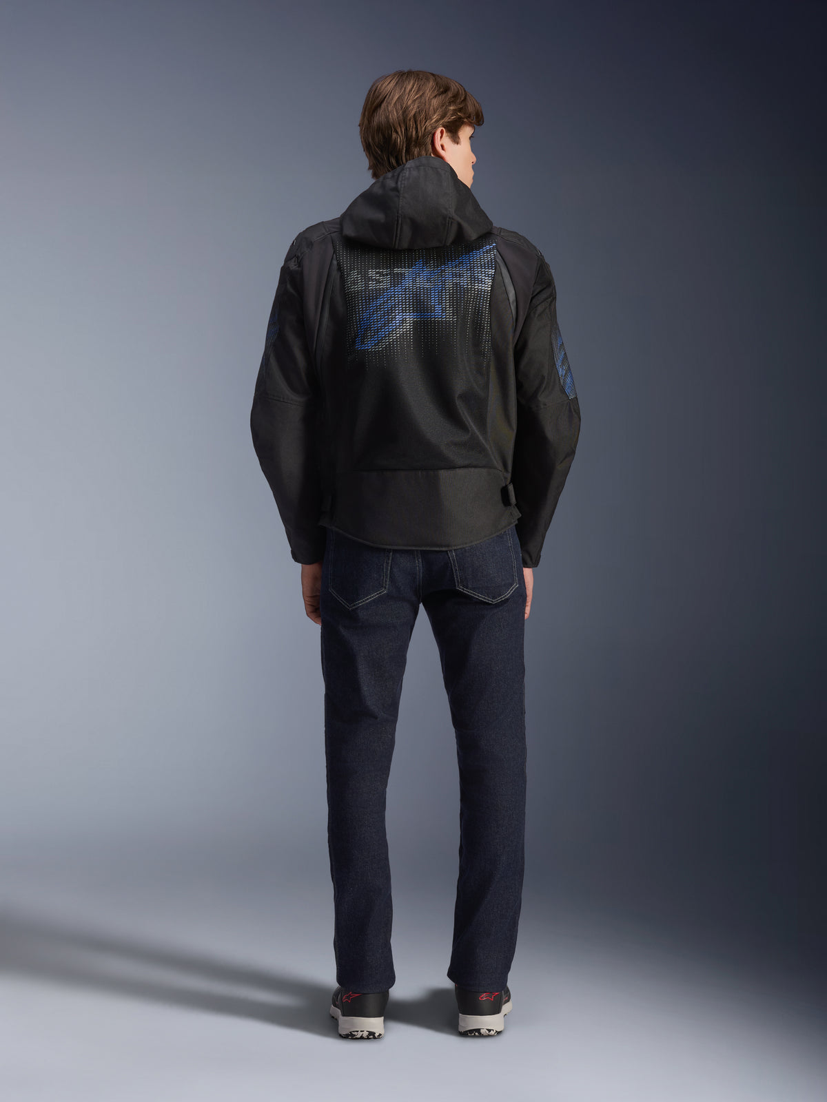 Alpinestars Zaca Air Venom WP Jacket, Black and Electric Blue, rear view featuring a large blue Alpinestars logo on the back mesh panel, detachable hood, worn with dark denim jeans and sneakers, sporty all-weather motorcycle gear.