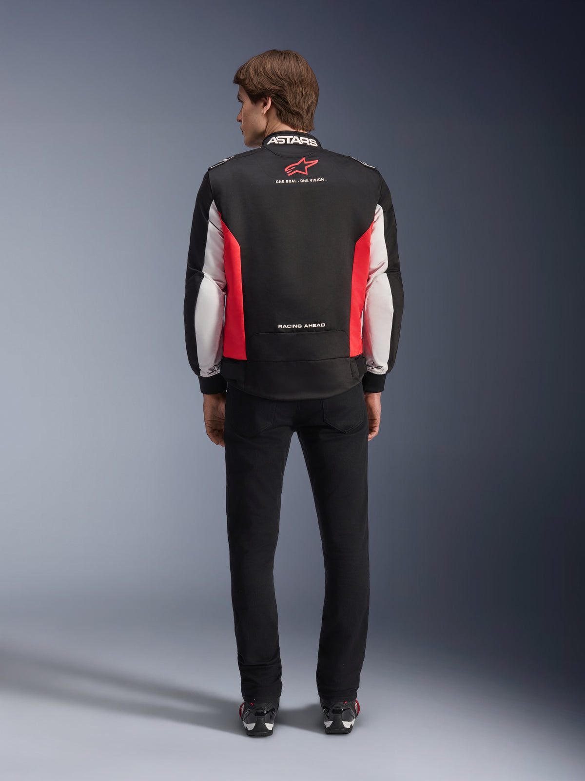 Alpinestars Monza Sport-jakke, tekstil-motorcykeljakke i sort, klar rød og hvid, set bagfra på en model, med rødt stjerne-logo og 'ASTARS'-branding på kraven med slogans, båret med sorte bukser og motorcykelstøvler.