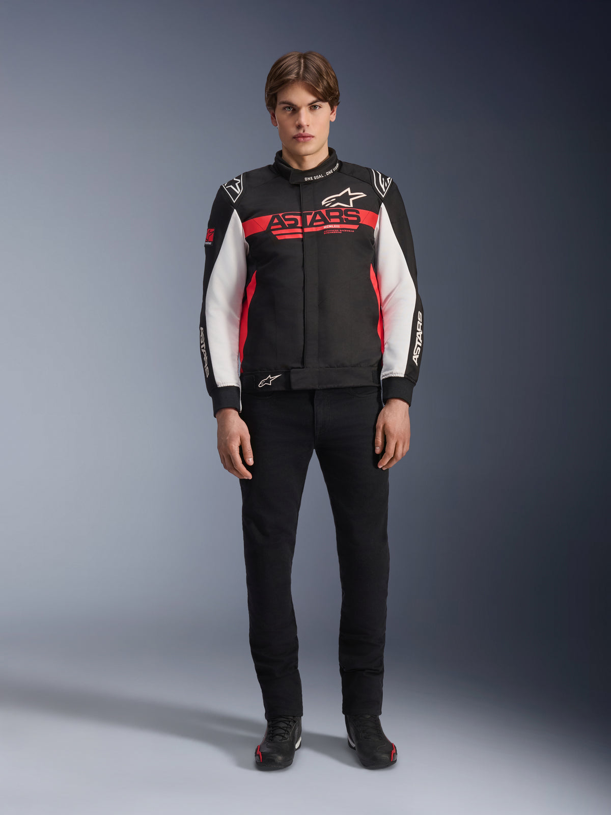 Mandlig model iført Alpinestars Monza Sport-jakke i sort, klar rød og hvid, med et stort rødt 'ASTARS'-logo over brystet og hvide paneler på ærmerne, kombineret med sorte bukser og sorte kørestøvler.