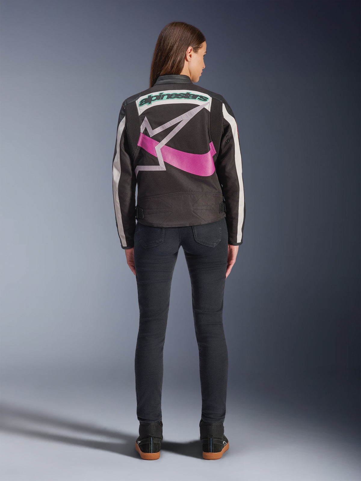 Rear view of a woman wearing the Alpinestars Idles Jacket, Black with off-white stripes, featuring a large purple and white Alpinestars star logo and green wordmark on the back, paired with dark slim-fit jeans, vintage-inspired urban motorcycle apparel