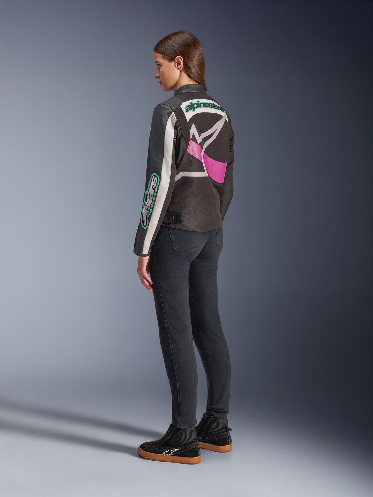 Alpinestars Idles Women’s Jacket, Motorcycle Jacket, Black Off White and Purple, viewed from the back on model, featuring a large wrap-around Alpinestars logo and vintage racing-inspired design, paired with dark jeans and black shoes.
