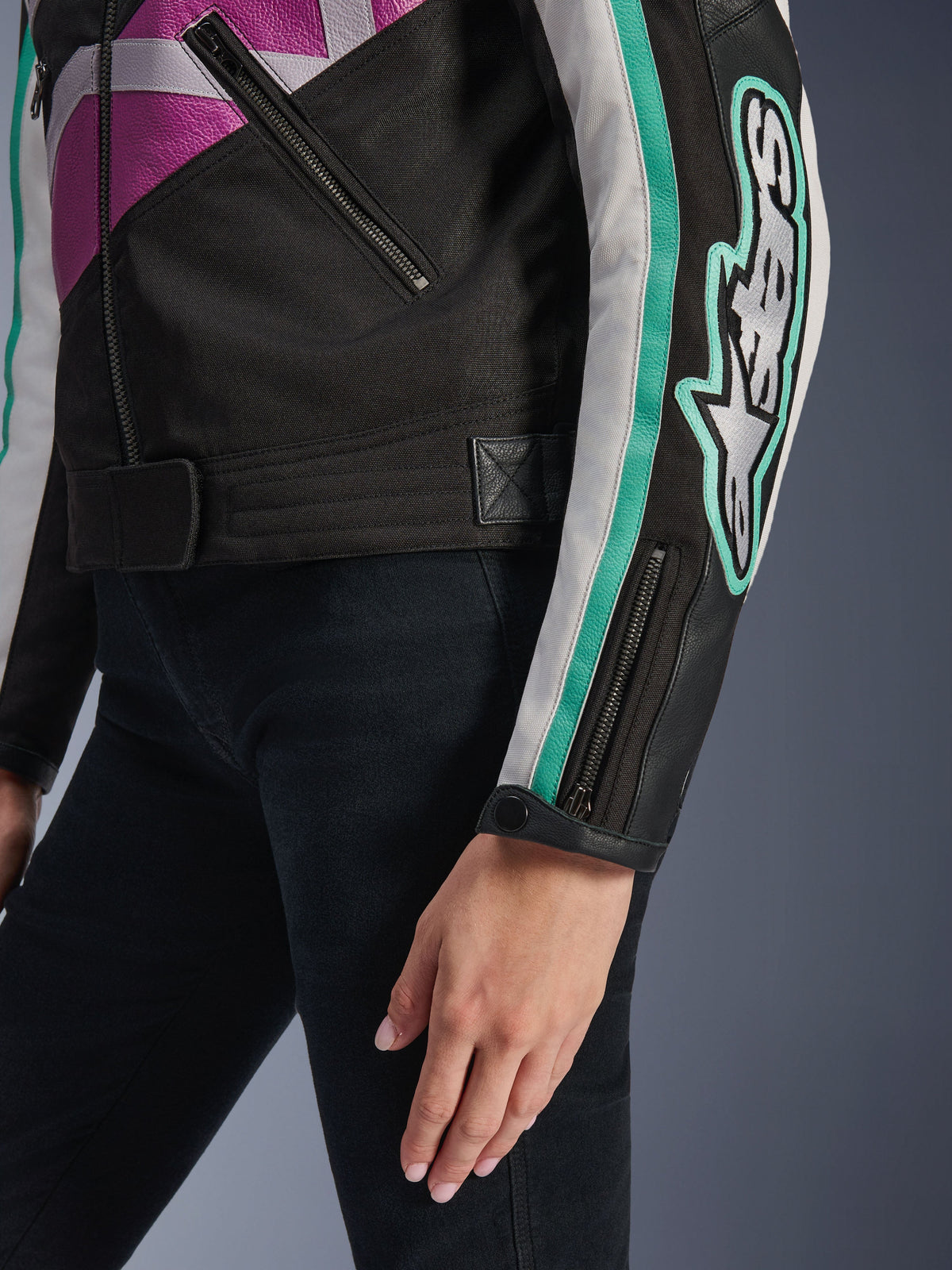 Alpinestars Idles Women's Jacket, close-up view of the sleeve and side torso, black with purple, white, and teal accents, featuring a bold Alpinestars logo on the arm, zippered cuff with snap, and waist adjustment strap, vintage-inspired motorcycle gear.