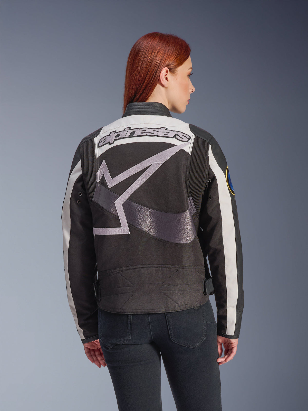 Alpinestars Idles Women’s Jacket, Black and Off White, back view featuring a large grey star logo and Alpinestars wordmark, worn by a red-haired model, vintage-inspired urban motorcycle jacket with accordion panels.