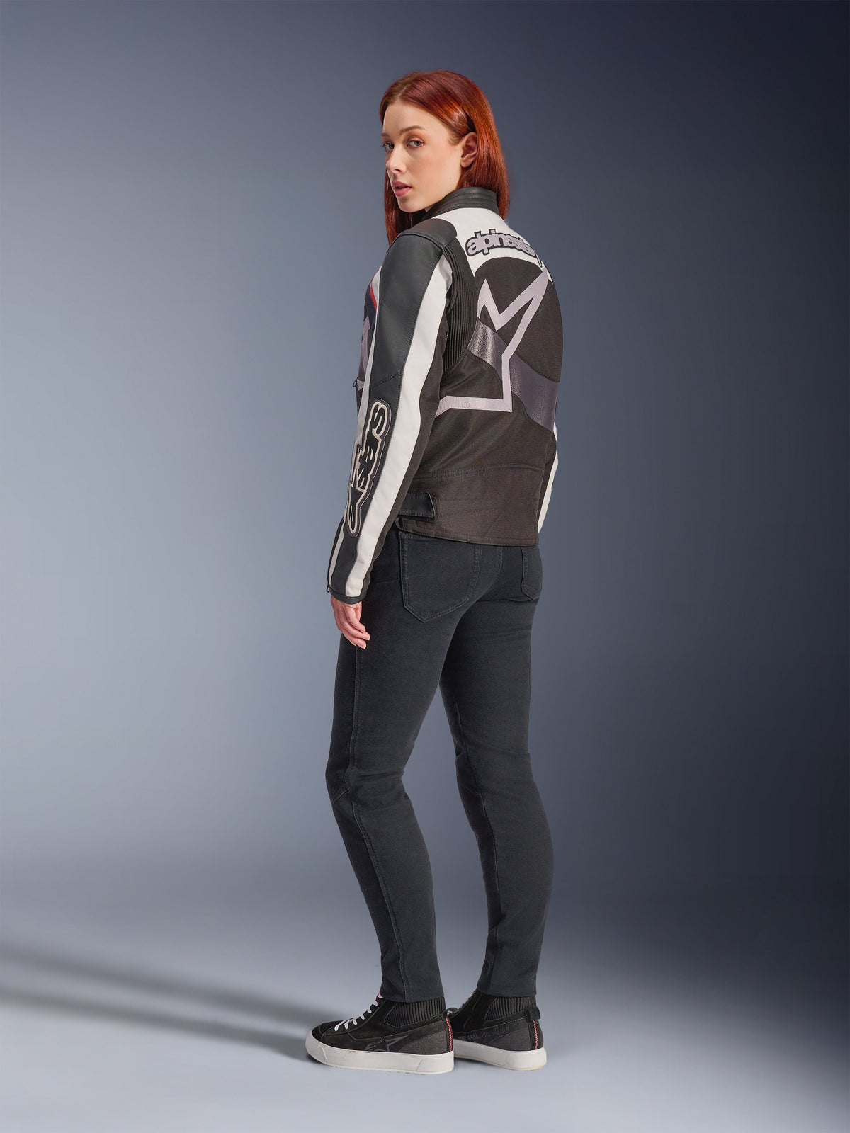 Woman wearing Alpinestars Idles Jacket, back view, Off White Flame Red and Black, featuring a large star logo on the back and Alpinestars wordmark on the sleeve, paired with black slim-fit pants and sneakers, urban motorcycle apparel for women