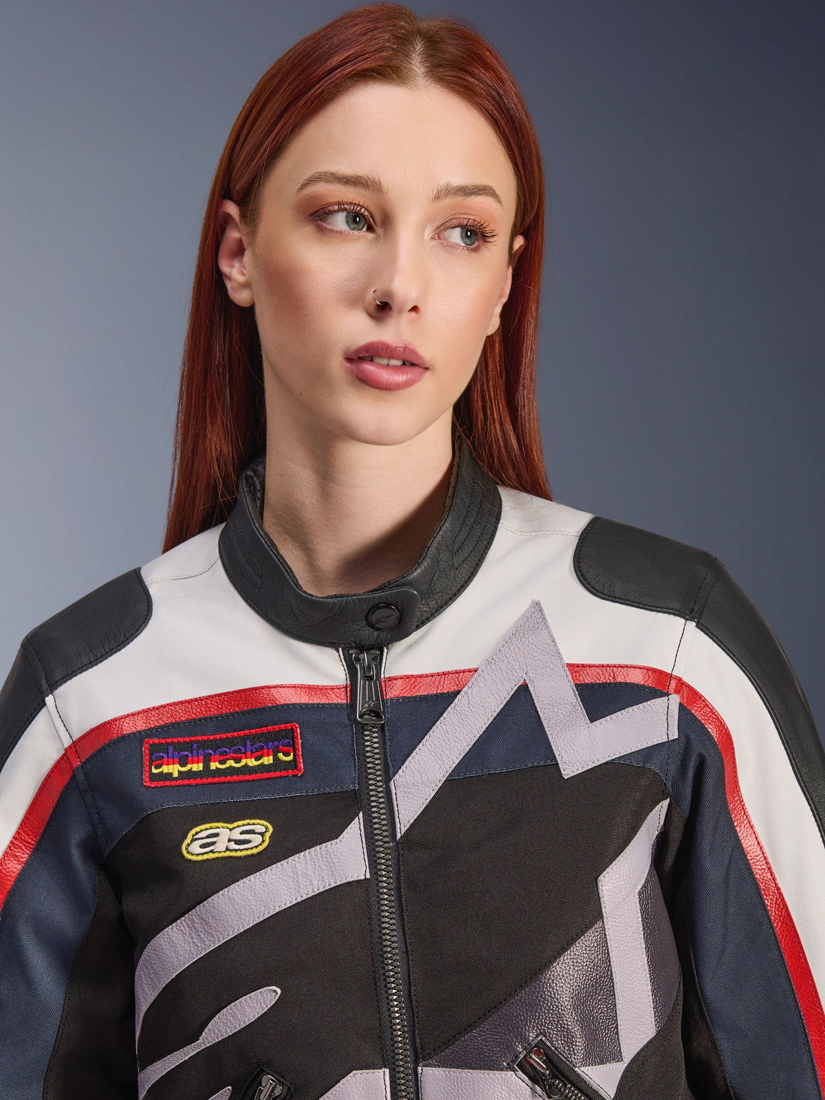Alpinestars Idles Women’s Jacket, close-up of a female model with red hair, Off White Flame Red and Black colorway, vintage-inspired racing design with logo patches and grey geometric star graphics, urban motorcycle riding gear.