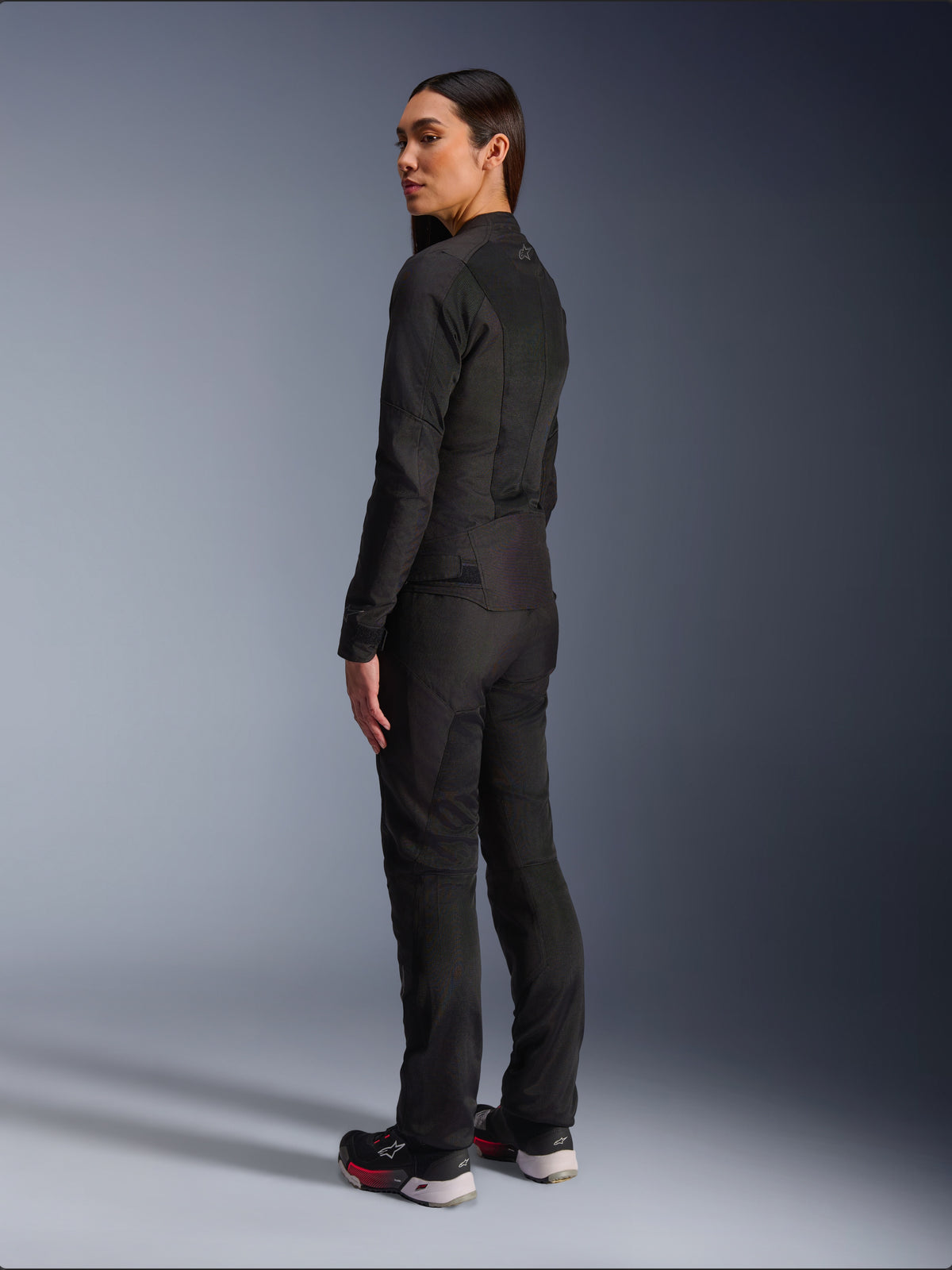 Kvinde iført Alpinestars Stella C-1 Air-jakke i sort, urban tekstil-motorcykeljakke med mesh-paneler, set bagfra fra siden der viser den slanke pasform og logoet øverst på ryggen, båret med matchende sorte bukser og sportssko, moderne motorcykeludstyr