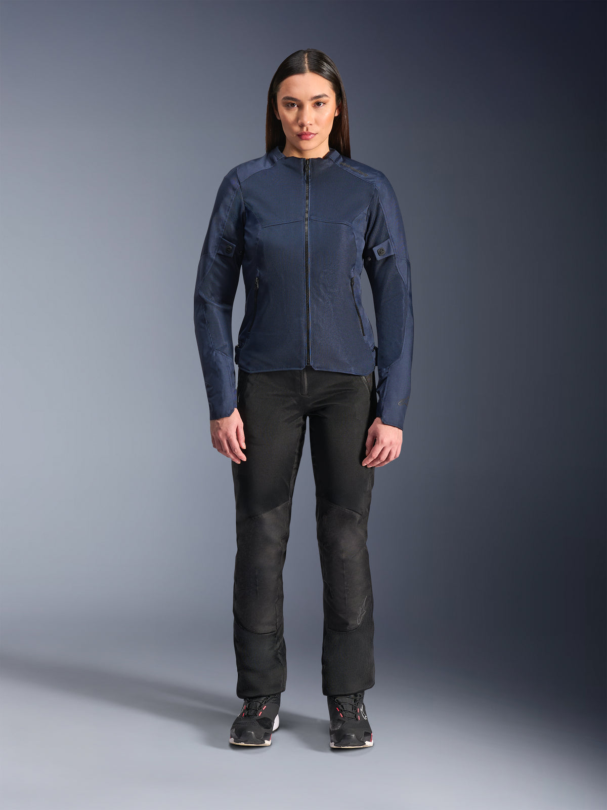 Kvindelig model iført Alpinestars Stella C-1 Air-jakke i blå, urban motorcykeljakke med tech mesh-paneler, kombineret med sorte motorcykelbukser og sorte sneakers, studiebillede set forfra