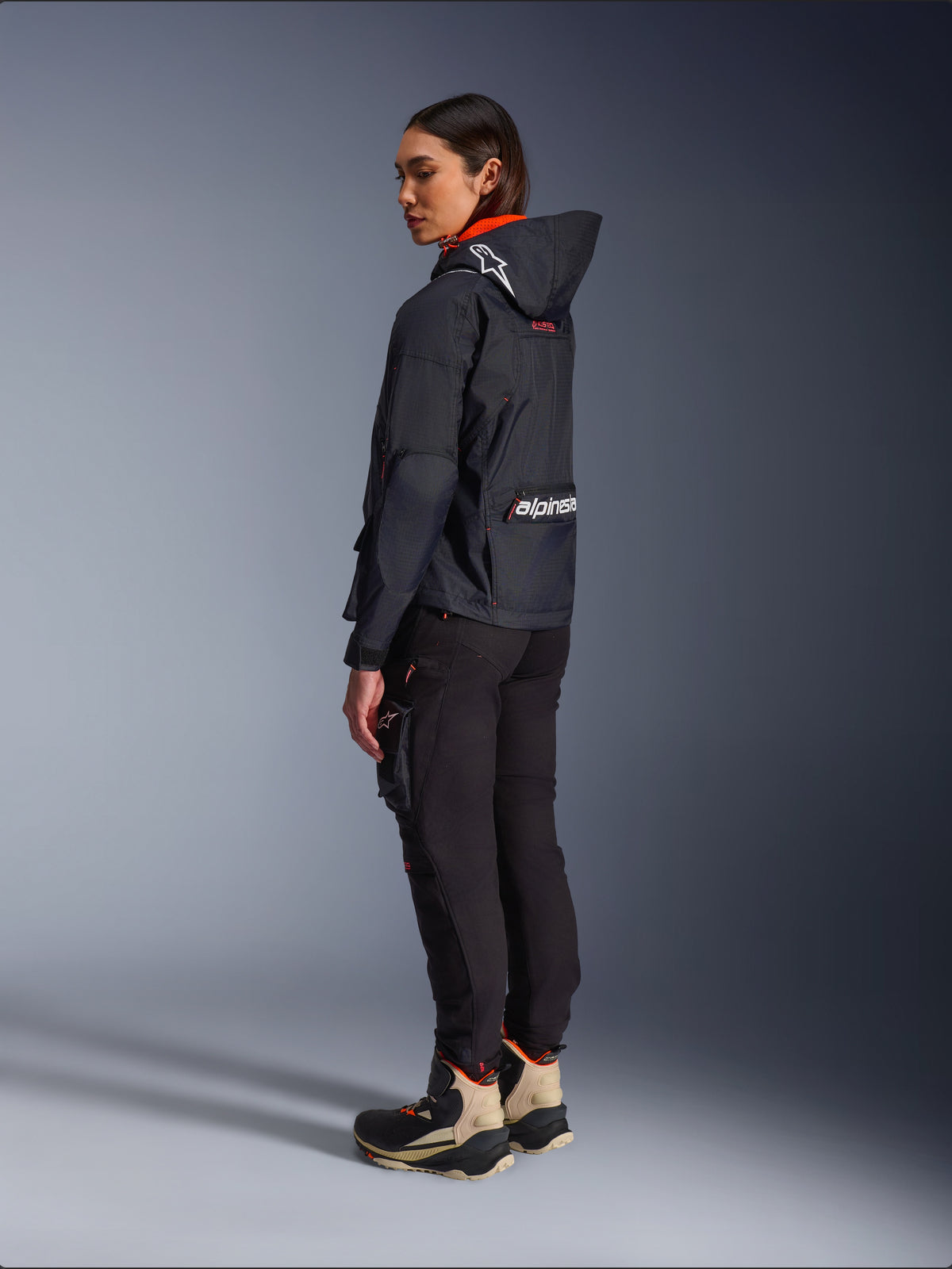 Alpinestars Actea Women's Pants, Black, modern motorcycle pants with a tapered fit and thigh cargo pocket, shown from a side-rear perspective on a model wearing a dark hooded jacket and riding boots, urban riding style.