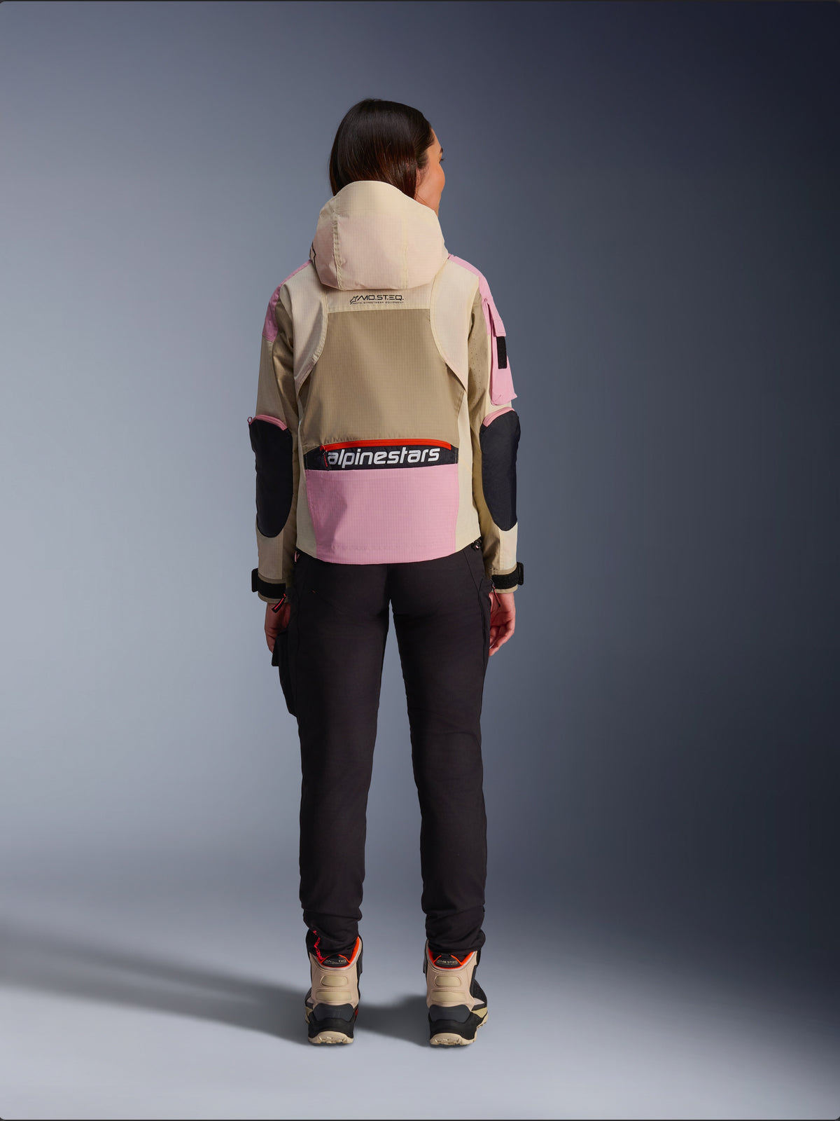 Kvindelig rytter set bagfra iført Alpinestars Molly Air-jakke i Sand Pink Almond, med beige hætte og lyserøde paneler med sorte detaljer, sorte ridebukser og beige motorcykelstøvler, urbant moderne motorcykelbeklædning