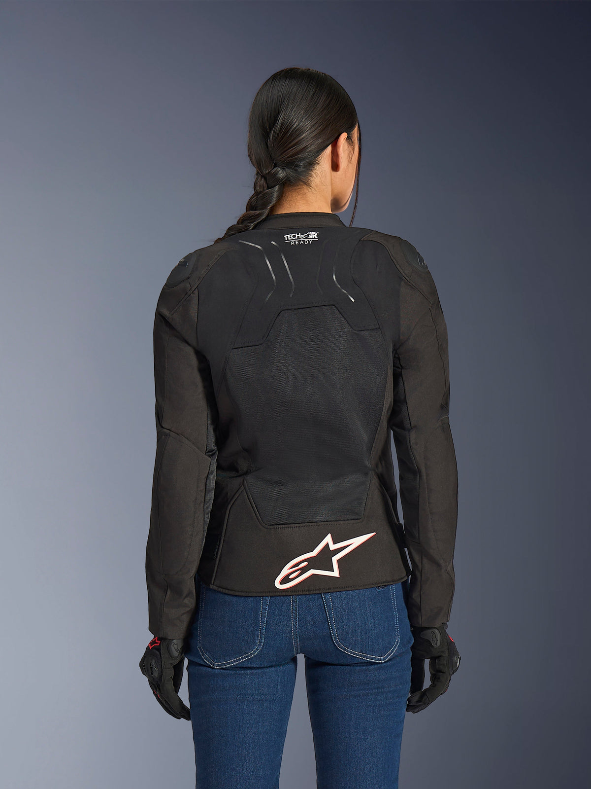 Alpinestars Stella SMX Air-jakke, set bagfra på kvindelig model, sort tekstil med high-flow mesh-paneler, 'TECH-AIR READY'-branding på den øvre ryg, stort hvidt Alpinestars-logo med rød kontur på den nederste del af ryggen, kombineret med blå denimjeans og sorte kørehandsker
