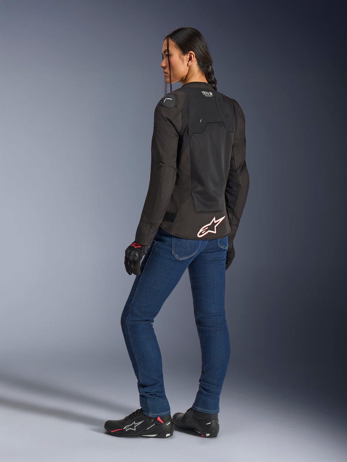 Alpinestars Stella SMX Air Jacket, Black with white and red logo, worn by a female rider seen from a rear three-quarter view, styled with blue motorcycle jeans and black riding footwear, featuring technical mesh construction for warm-weather sport riding.