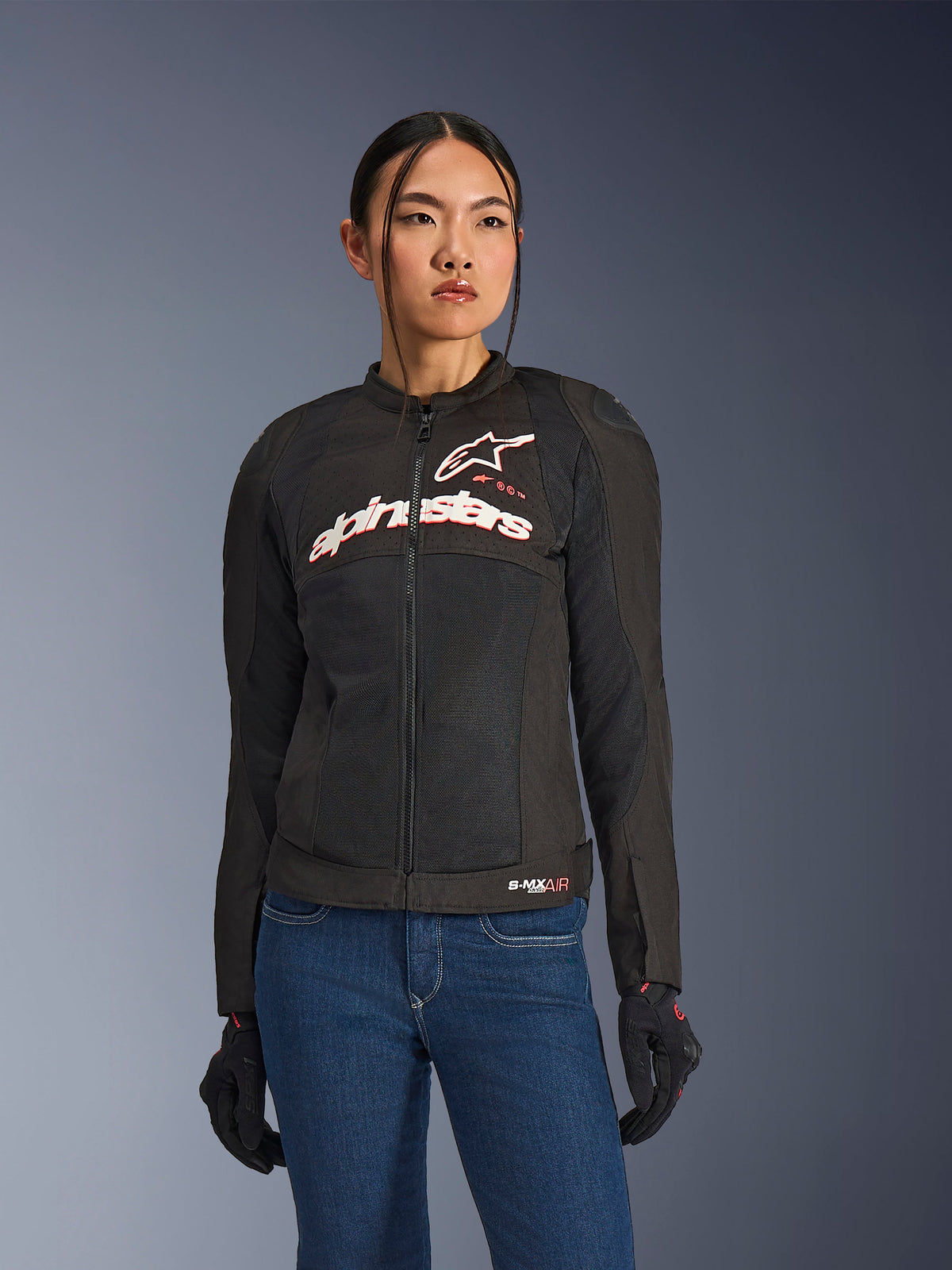 Woman wearing Alpinestars Stella SMX Air Jacket, Black White Red Fluo, textile motorcycle jacket with ventilated mesh and white chest logo, styled with blue jeans and black riding gloves, feminine fit for sport riding