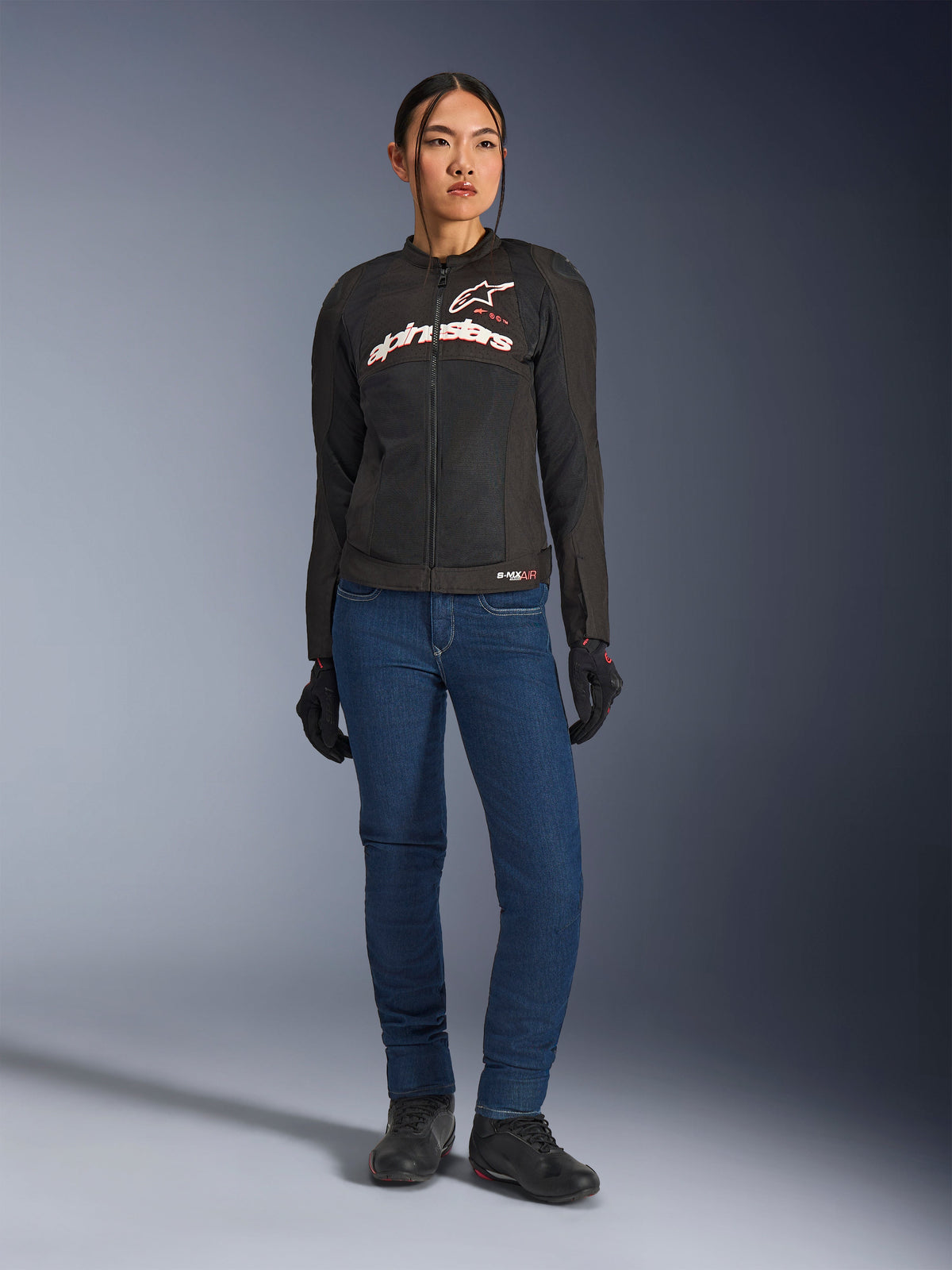 Female rider wearing the Alpinestars Stella SMX Air Jacket, Black with White and Red Fluo branding, paired with blue denim riding pants and black gloves, sporty ventilated textile jacket for road riding