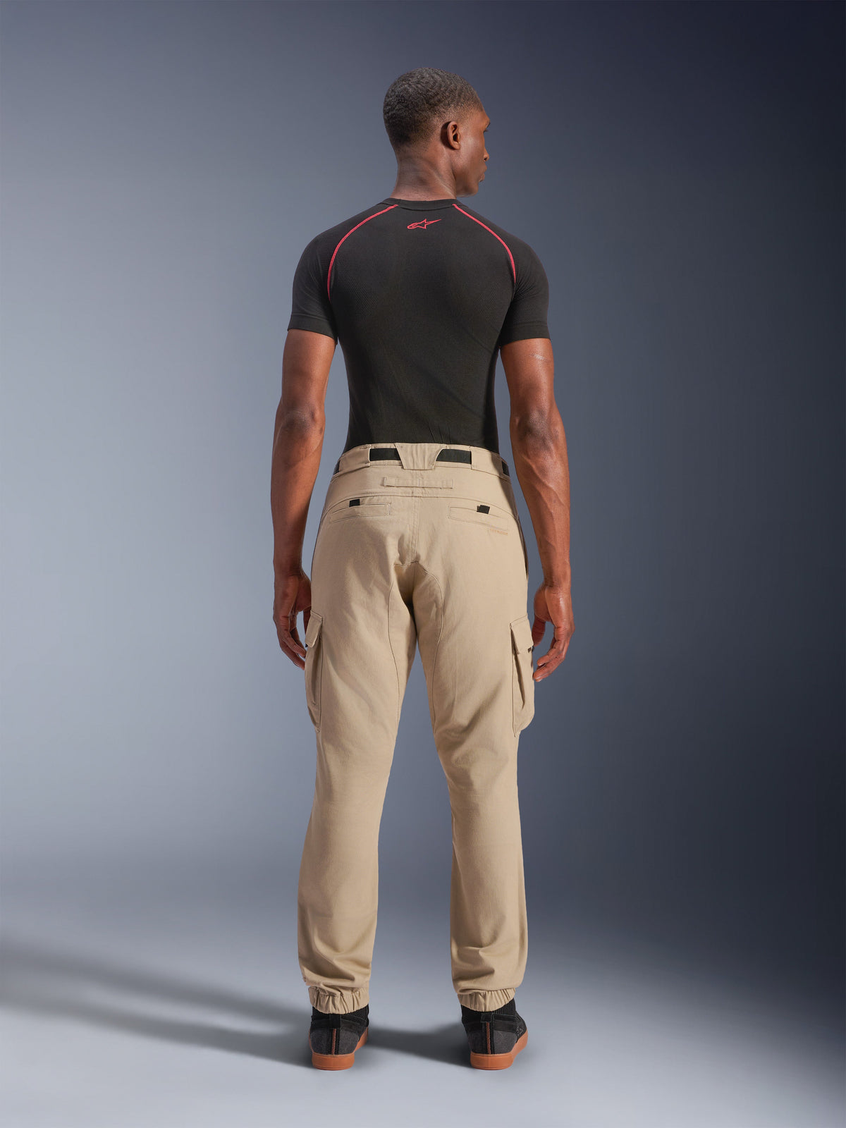 Alpinestars Flex-AST Cargo Pants - Short, lys beige, set bagfra på en model, regular tapered fit med elastiske sømme og cargolommer, båret med et sort teknisk basislag med røde detaljer, moderne urbant motorcykeltøj