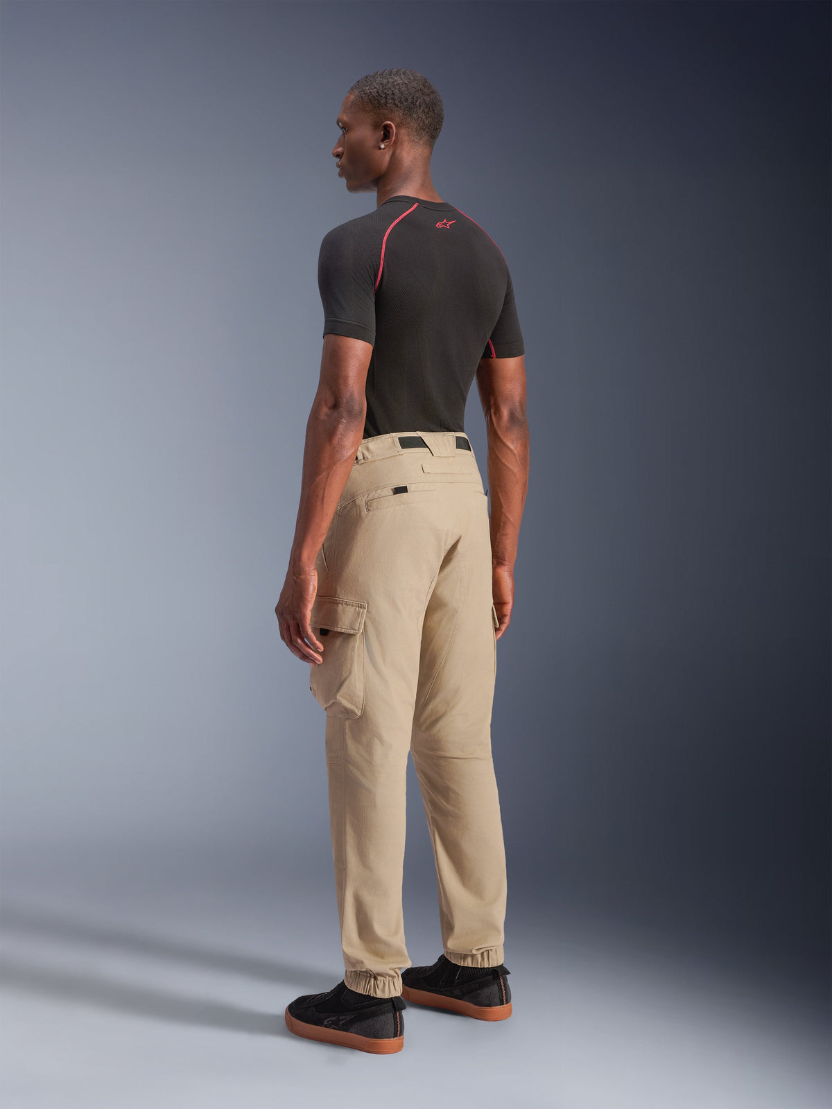 Alpinestars Flex-AST Cargo bukser - Short, urbane motorcykelbukser i lys beige, set bagfra fra siden, viser cargolommer i siden og elastiske opslag, stylet med en sort kortærmet overdel og sorte hverdagssko