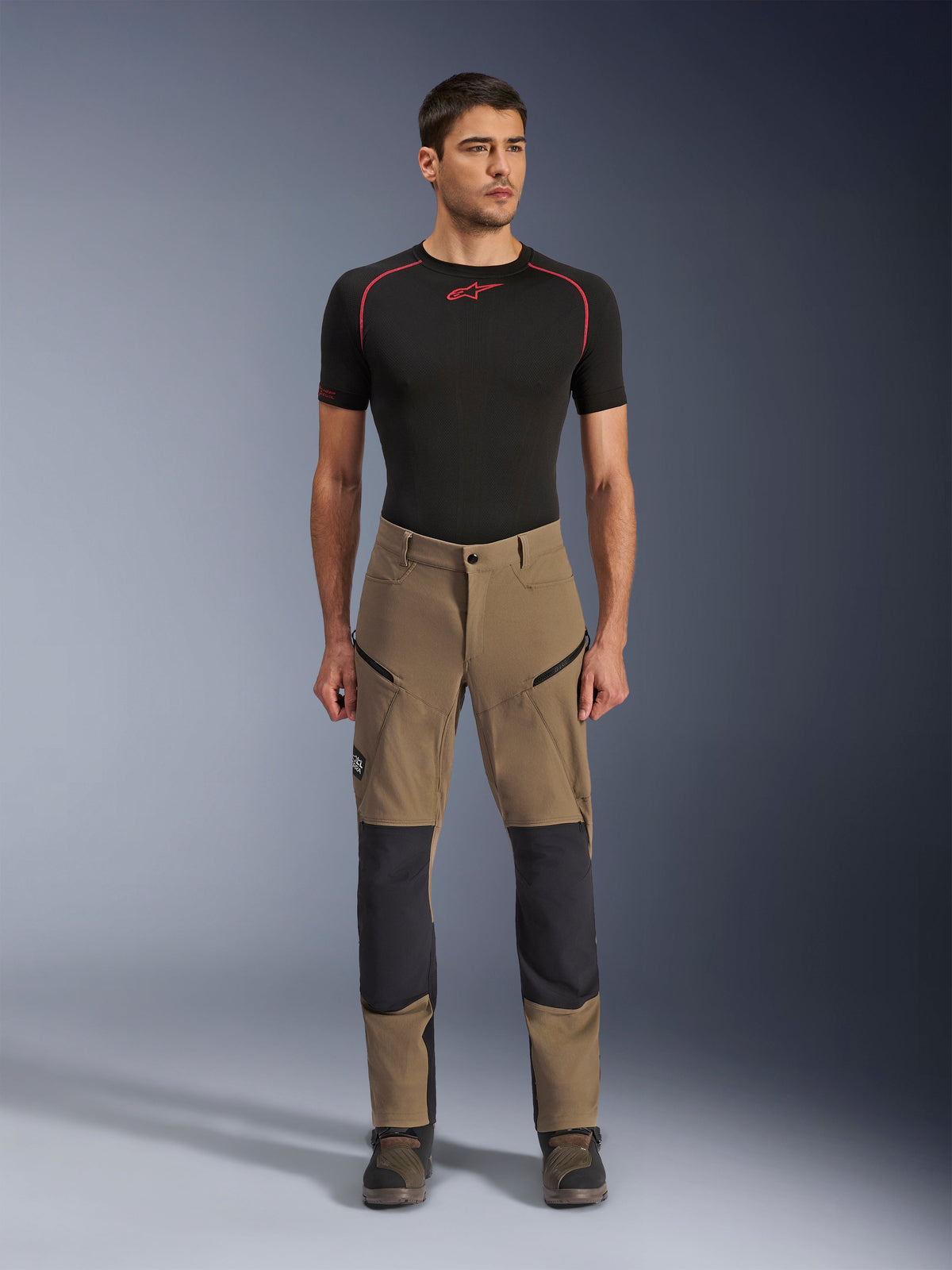 Alpinestars Flex-AST Explorer Pants, Adventure Motorcycle Pants, Walnut Black, worn by a model with a black base layer and boots, featuring tan stretch textile with black reinforced knee panels and zippered cargo pockets, designed for adventure touring.